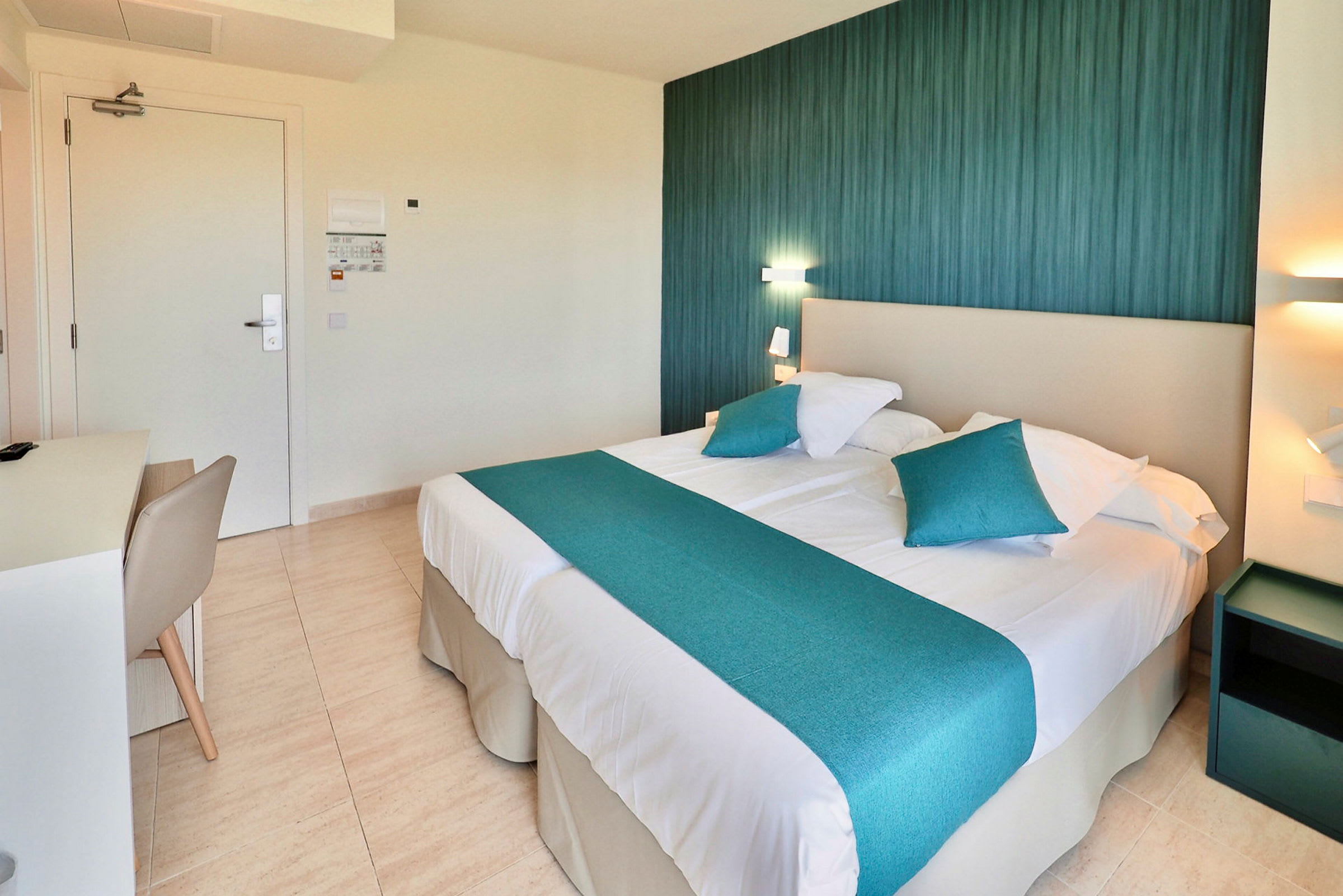 Ett hotellrum med en dubbelsäng som har blå dekorativa kuddar och sängöverkast. En lampa och ett nattduksbord syns bredvid sängen. (AI-genererad text)