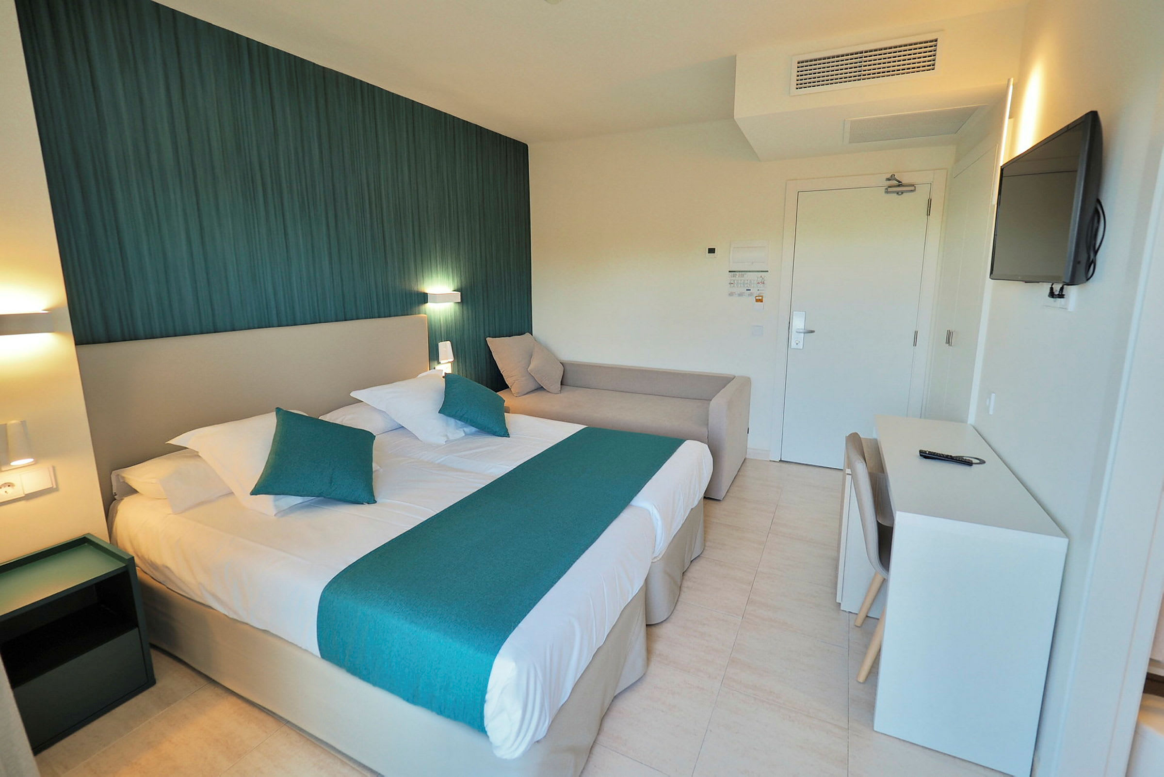 Ett hotellrum med en dubbelsäng, en soffa och ett skrivbord. Väggarna är ljusa med en mörkare accentvägg bakom sängen. (AI-genererad text)