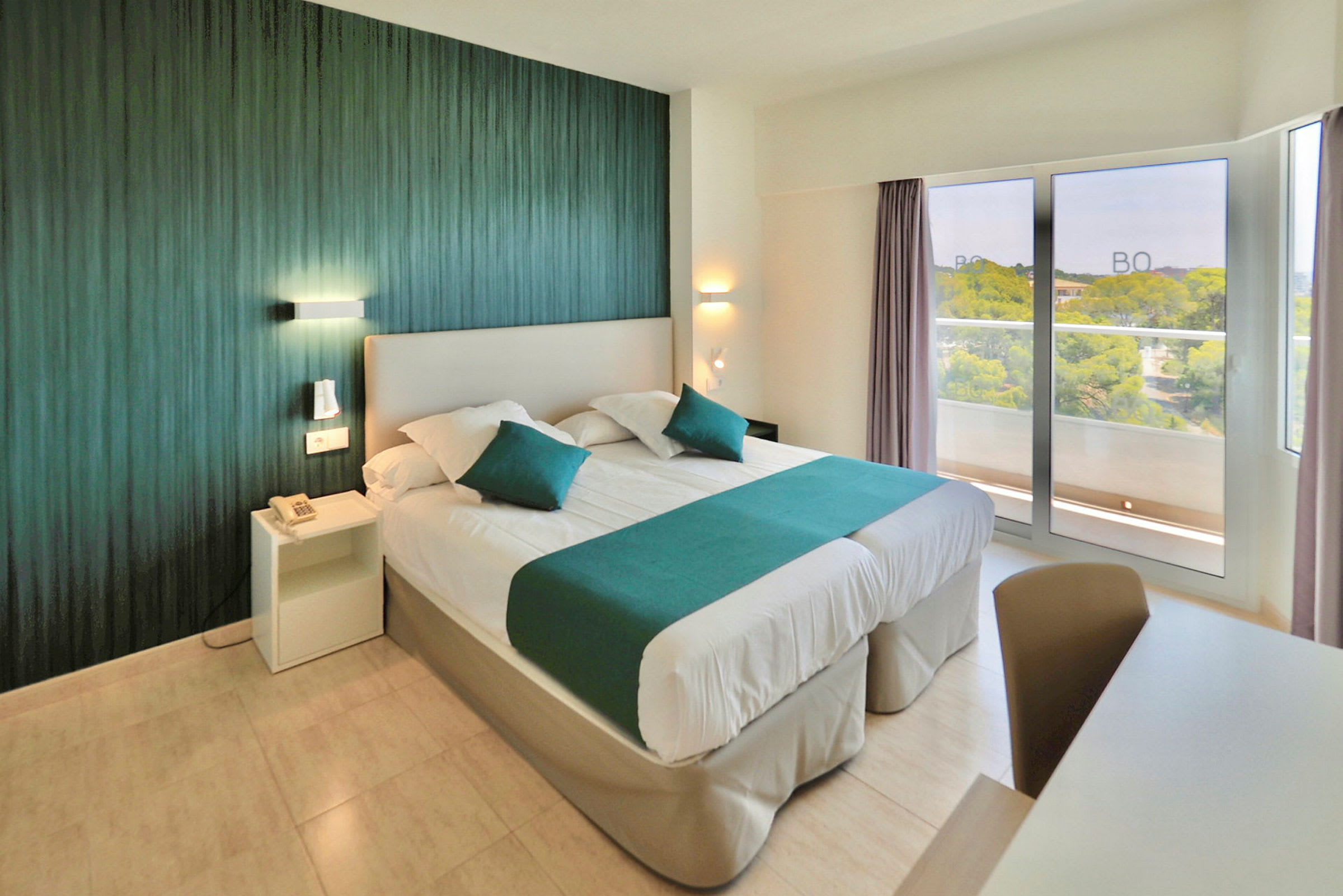 Ett hotellrum med en dubbelsäng bäddad med vita sängkläder och gröna detaljer. En modern inredning med en grön vägg och stora fönster som vetter mot balkongen. (AI-genererad text)