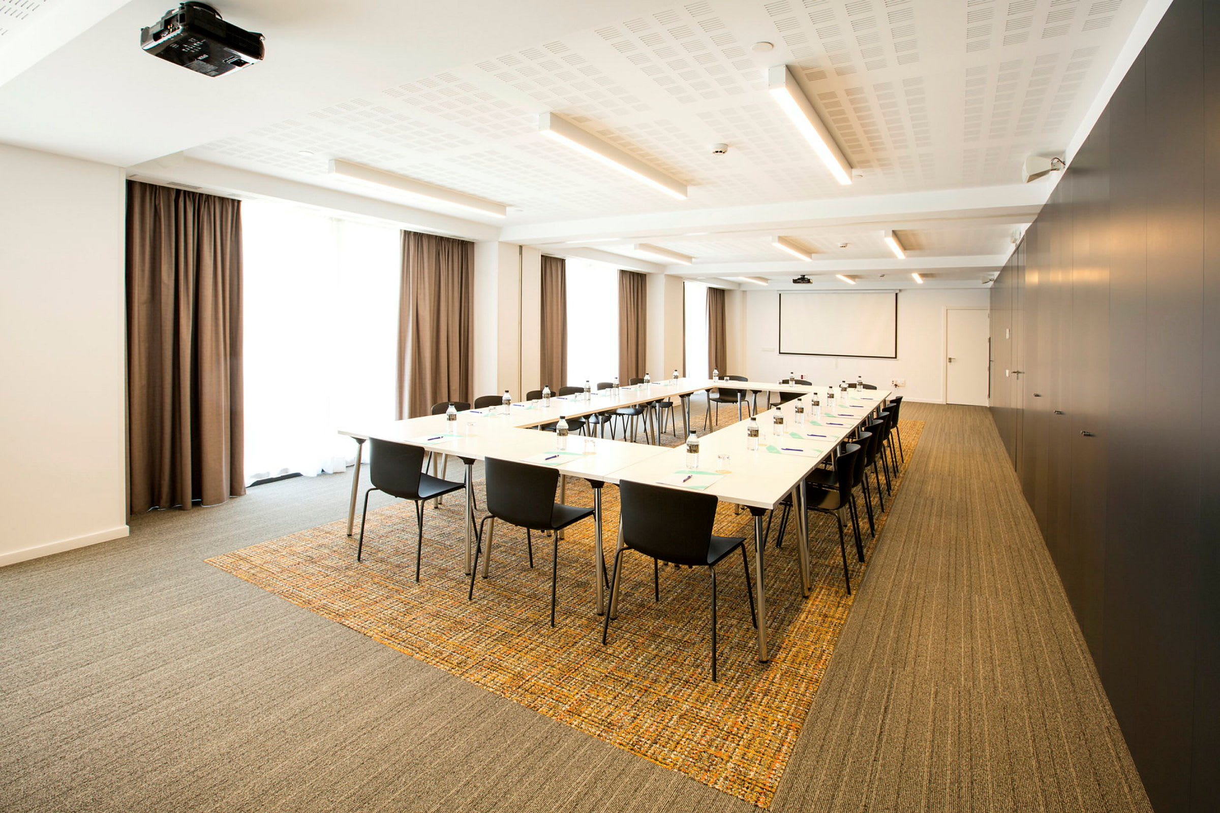 Konferensrum med bord arrangerade i U-form, omgivna av svartklädda stolar. Fönster med gardiner syns i bakgrunden. (AI-genererad text)