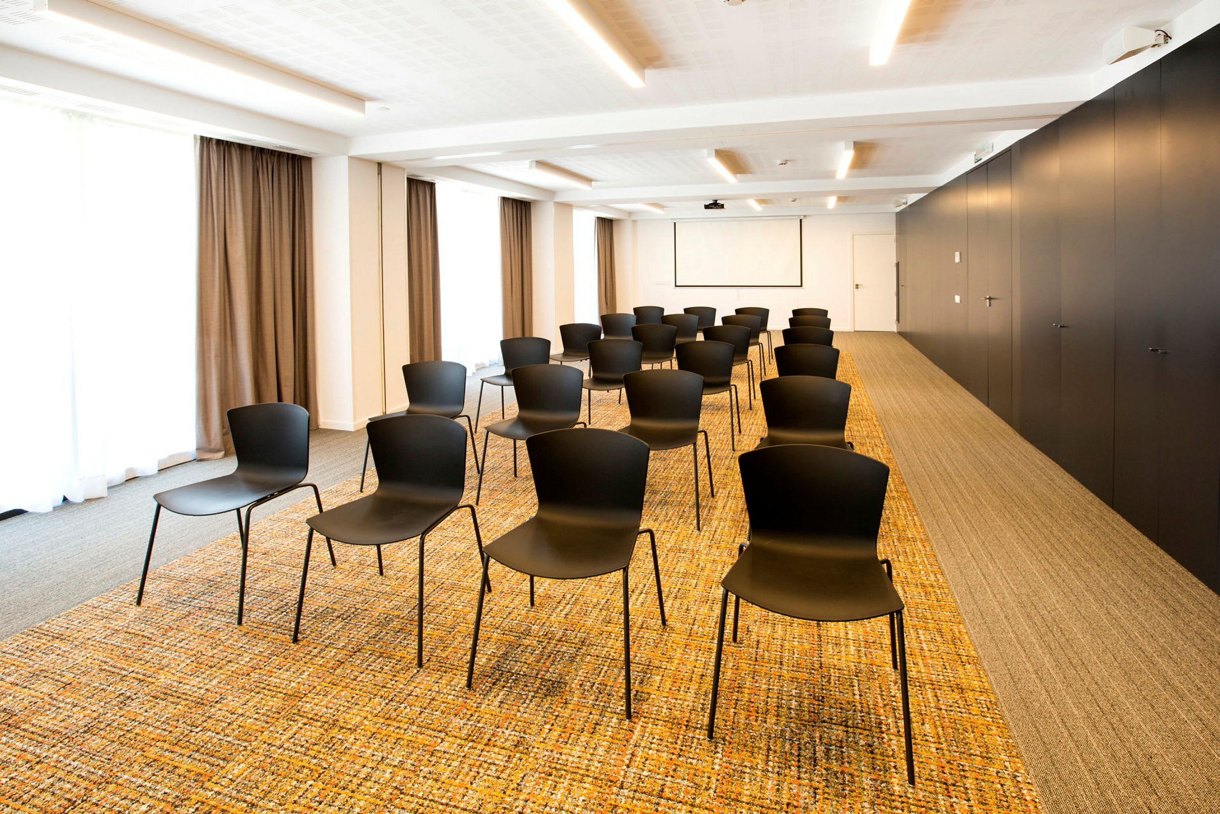 Ett konferensrum med svarta stolar i rader på en mönstrad matta. Det finns en projektorduk på väggen och draperier vid fönstren. (AI-genererad text)