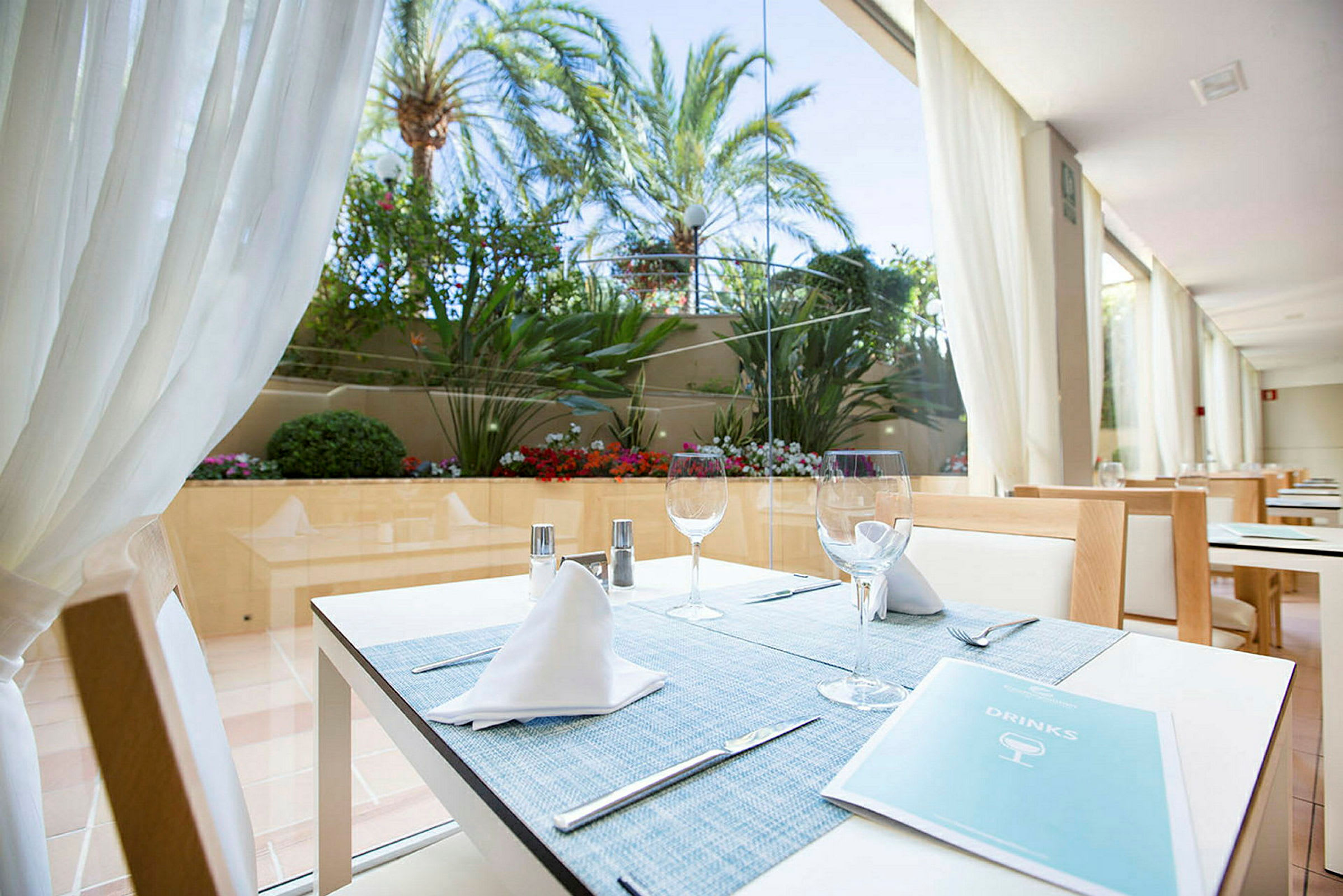 En restaurang med ett dukat bord nära ett stort fönster som vetter mot en trädgård med palmer och blomster. (AI-genererad text)
