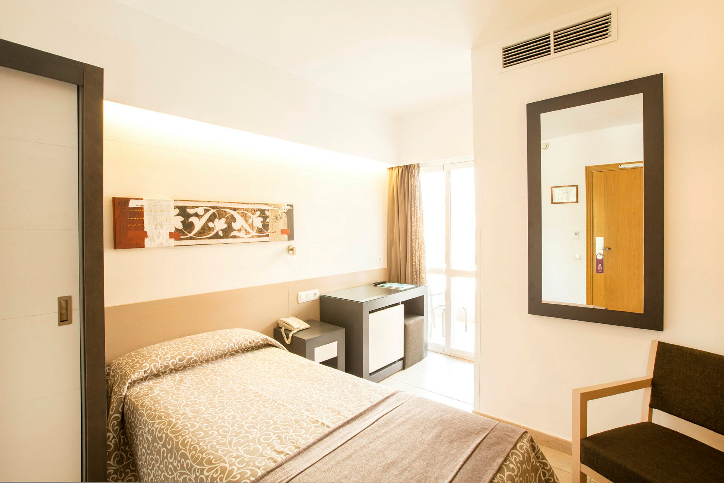 Hotellrum med en enkel säng klädd i ett mönstrat överkast, ett skrivbord med en stol, och en spegel på väggen. En glasad dörr leder ut till en balkong. (AI-genererad text)