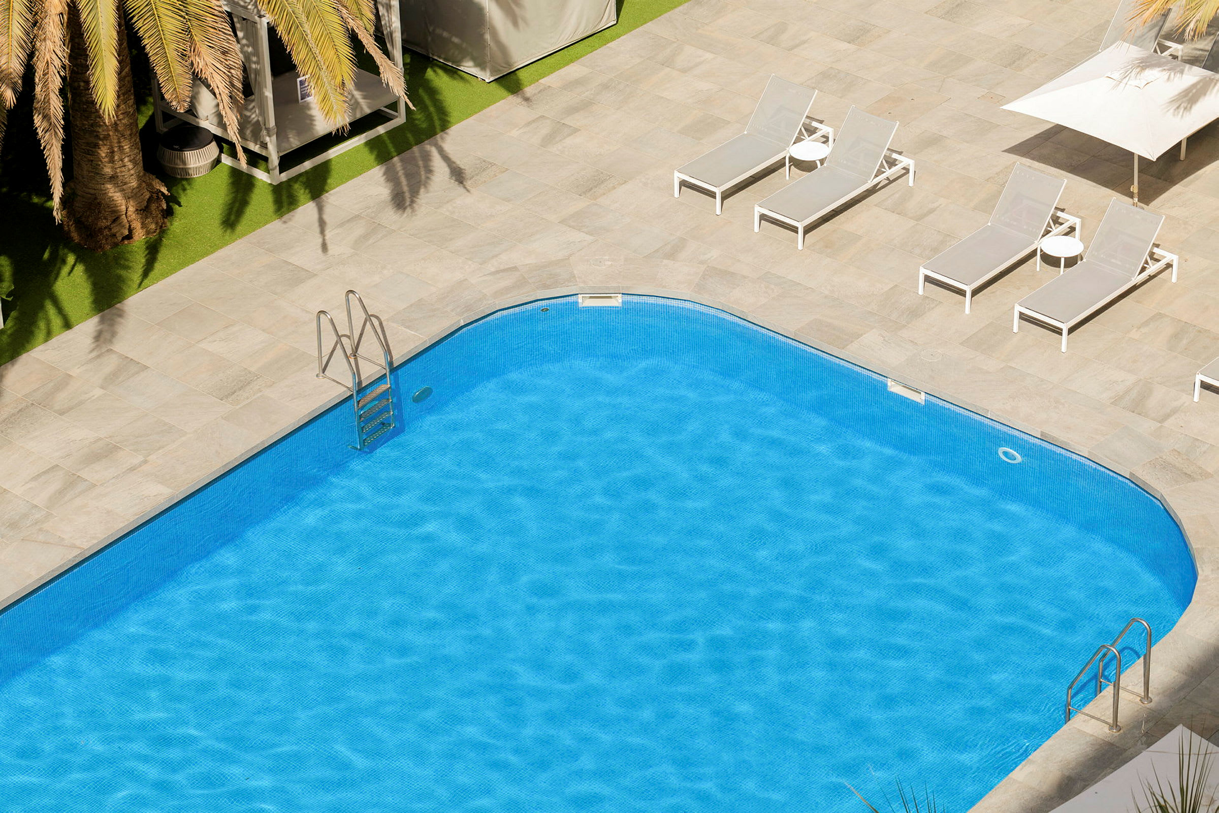 En swimmingpool med klart blått vatten och omgiven av solstolar och parasoller på en stenlagd yta. (AI-genererad text)