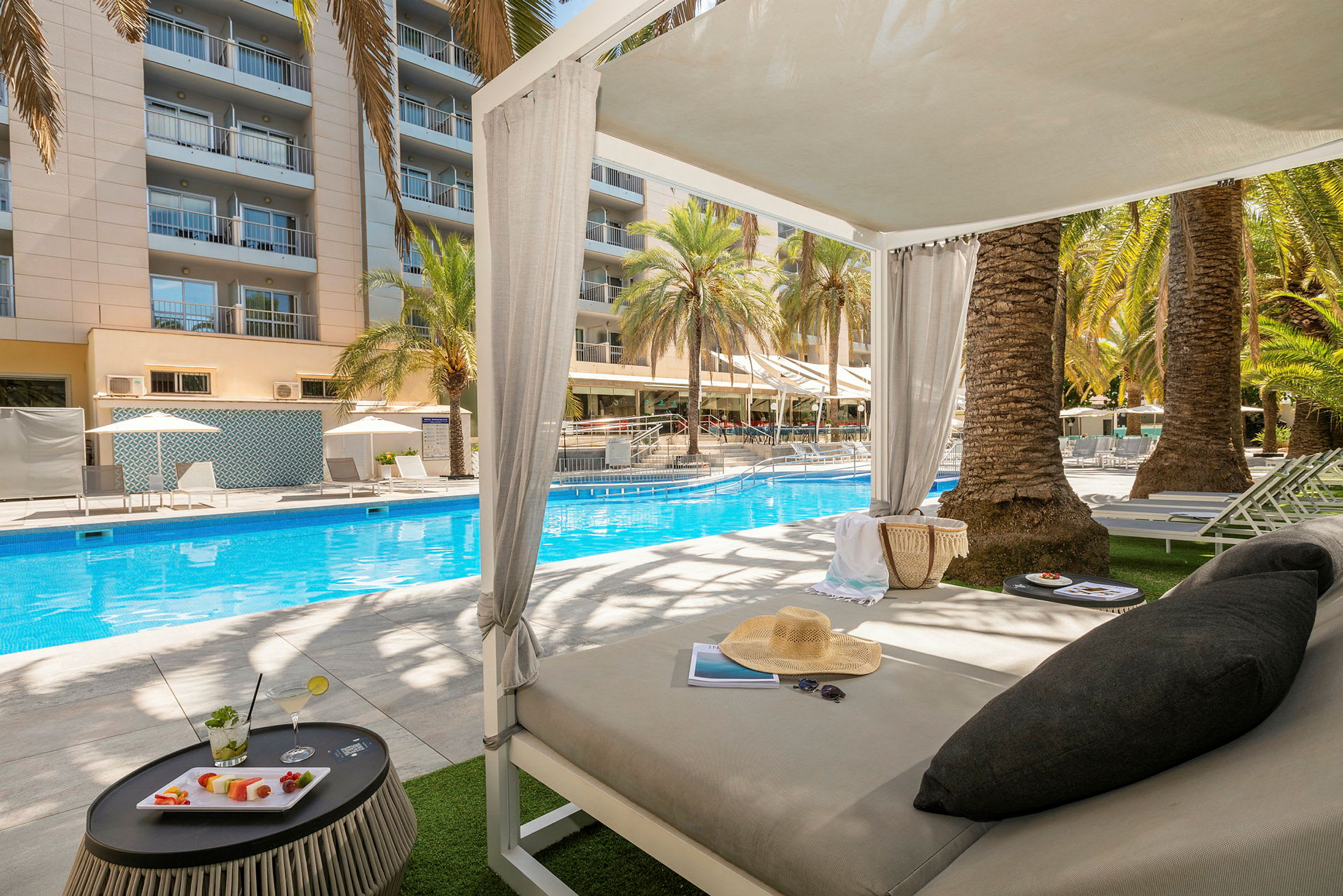 En bild av ett hotellområde med en pool omgivet av solstolar och parasoller. I förgrunden finns en solbädd under ett skuggande tak. (AI-genererad text)