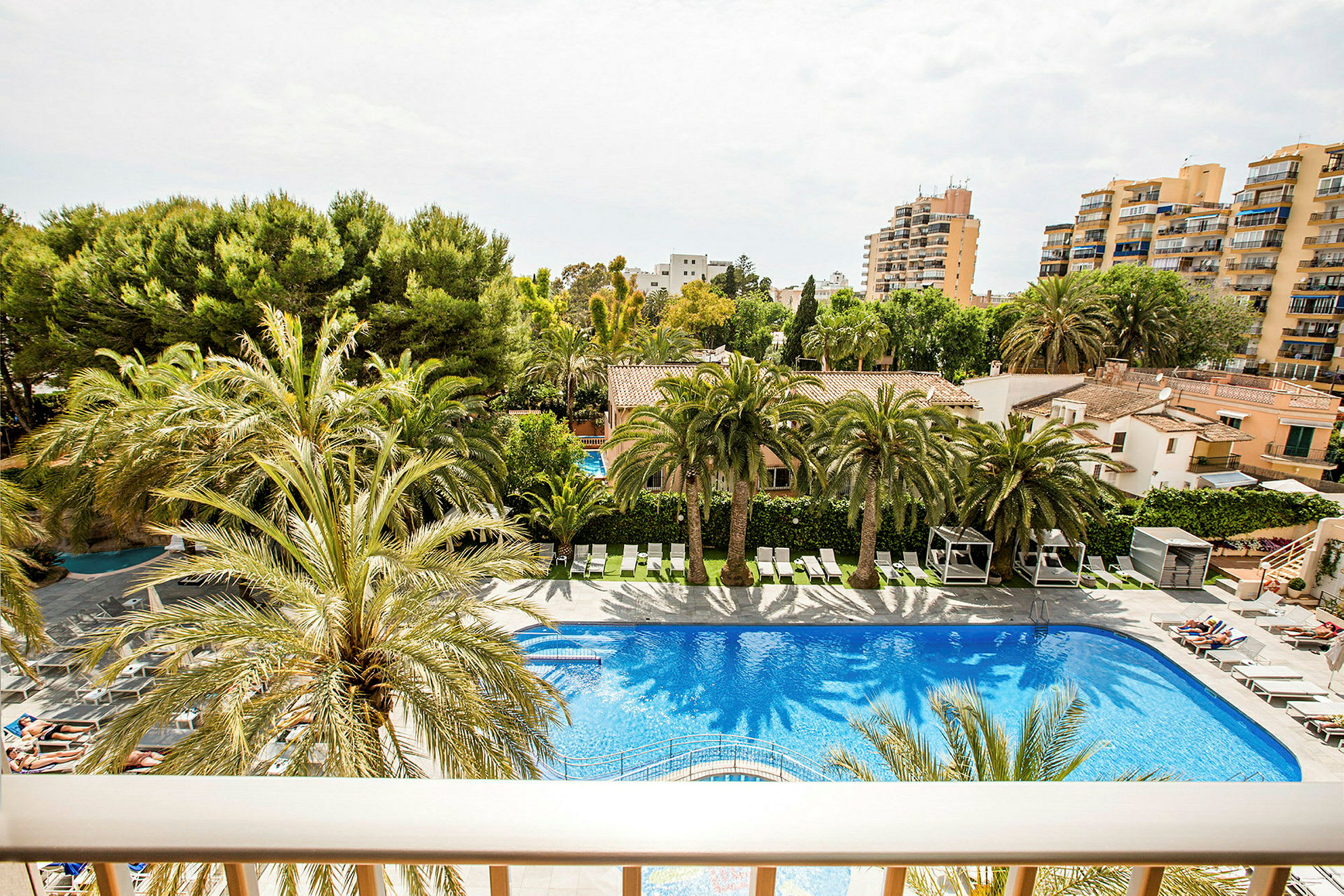 En bild av en pool vid ett hotellområde omgiven av palmer med byggnader i bakgrunden. (AI-genererad text)