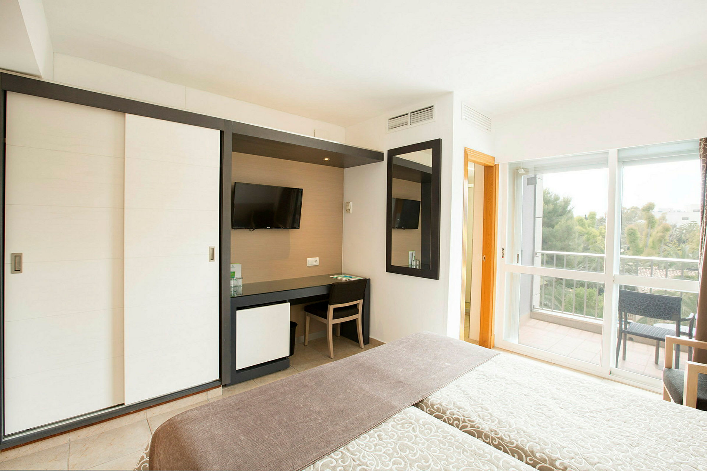 Ett hotellrum med en säng, en garderob och ett skrivbord framför en TV. Rummet har stora fönster med utsikt över en balkong. (AI-genererad text)