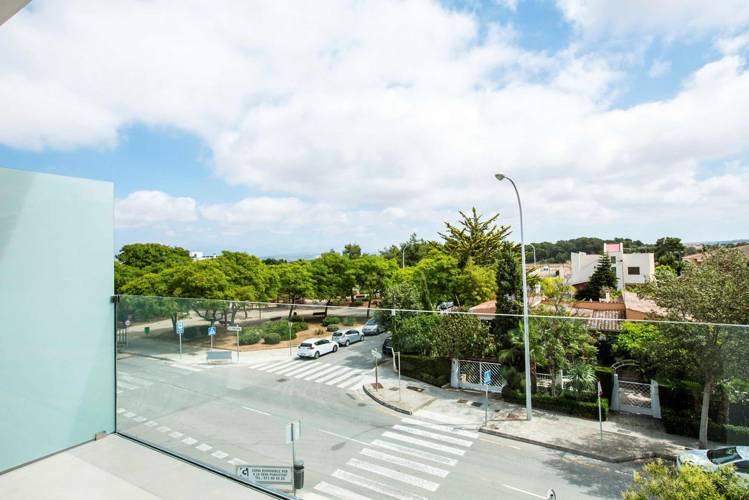 Utsikt från en balkong med glasräcke över en stadsgata och grönområden, med park och hus i bakgrunden. (AI-genererad text)