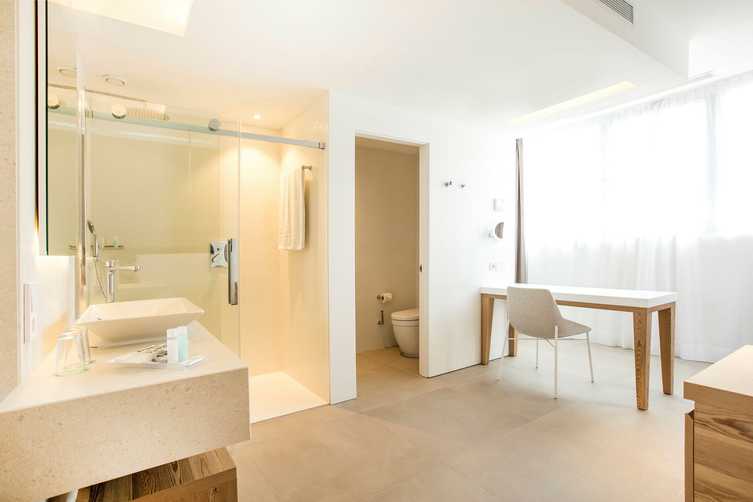 Ett modernt hotellrum med badrum i ljusa nyanser. Bilden visar ett tvättställ, en dusch med glasdörr, en toalett och en arbetsplats med skrivbord och stol. (AI-genererad text)