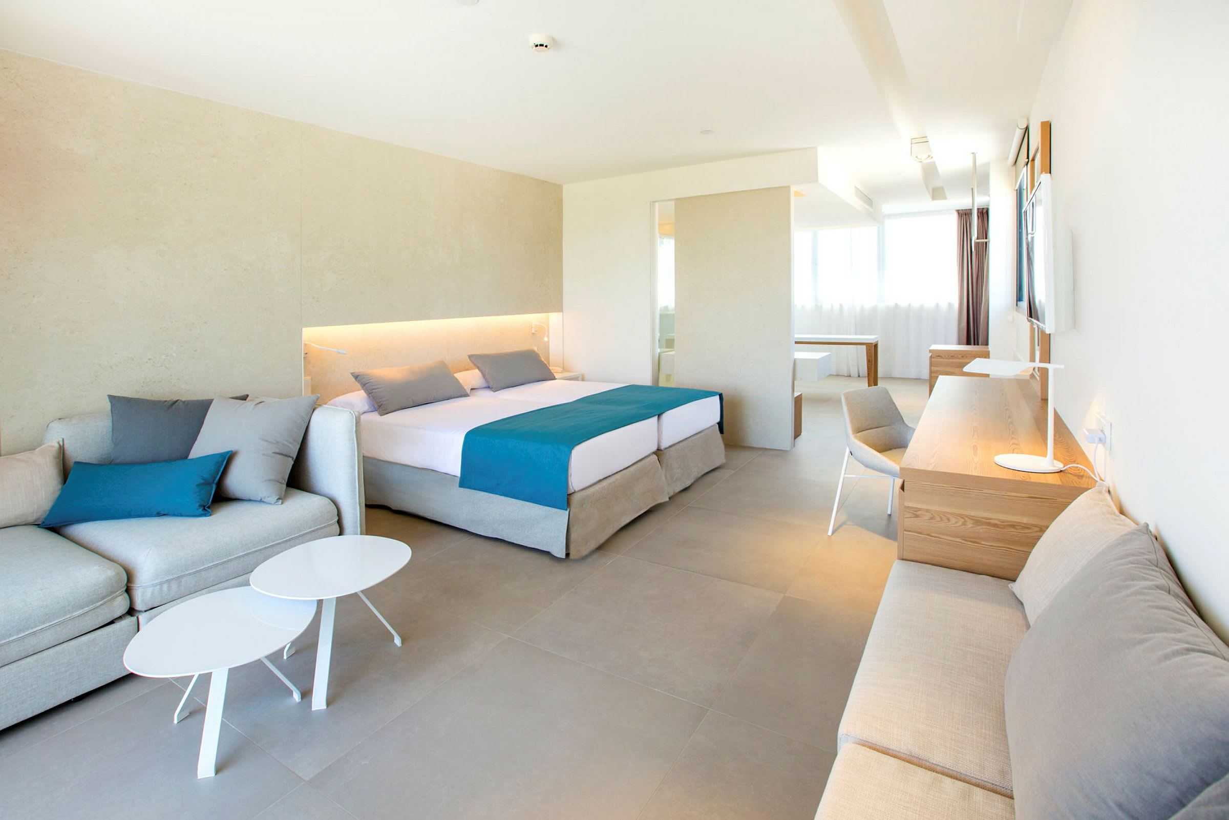 Ett ljust och modernt hotellrum med en stor säng, sittplats, skrivbord och garderob, med neutrala färger och blå accenter. (AI-genererad text)