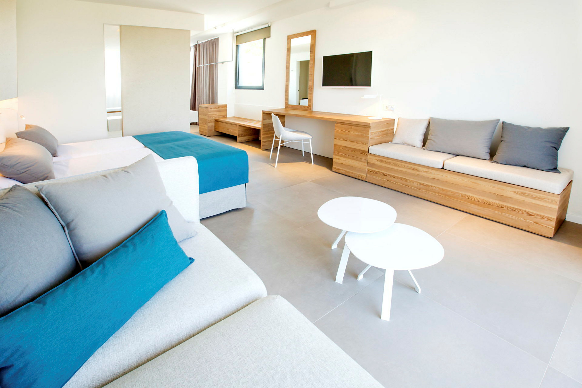 Ett modernt hotellrum med en stor säng, en soffa, ett skrivbord och en väggmonterad TV. Möblerna är träfärgade och rummet har en minimalistisk design. (AI-genererad text)