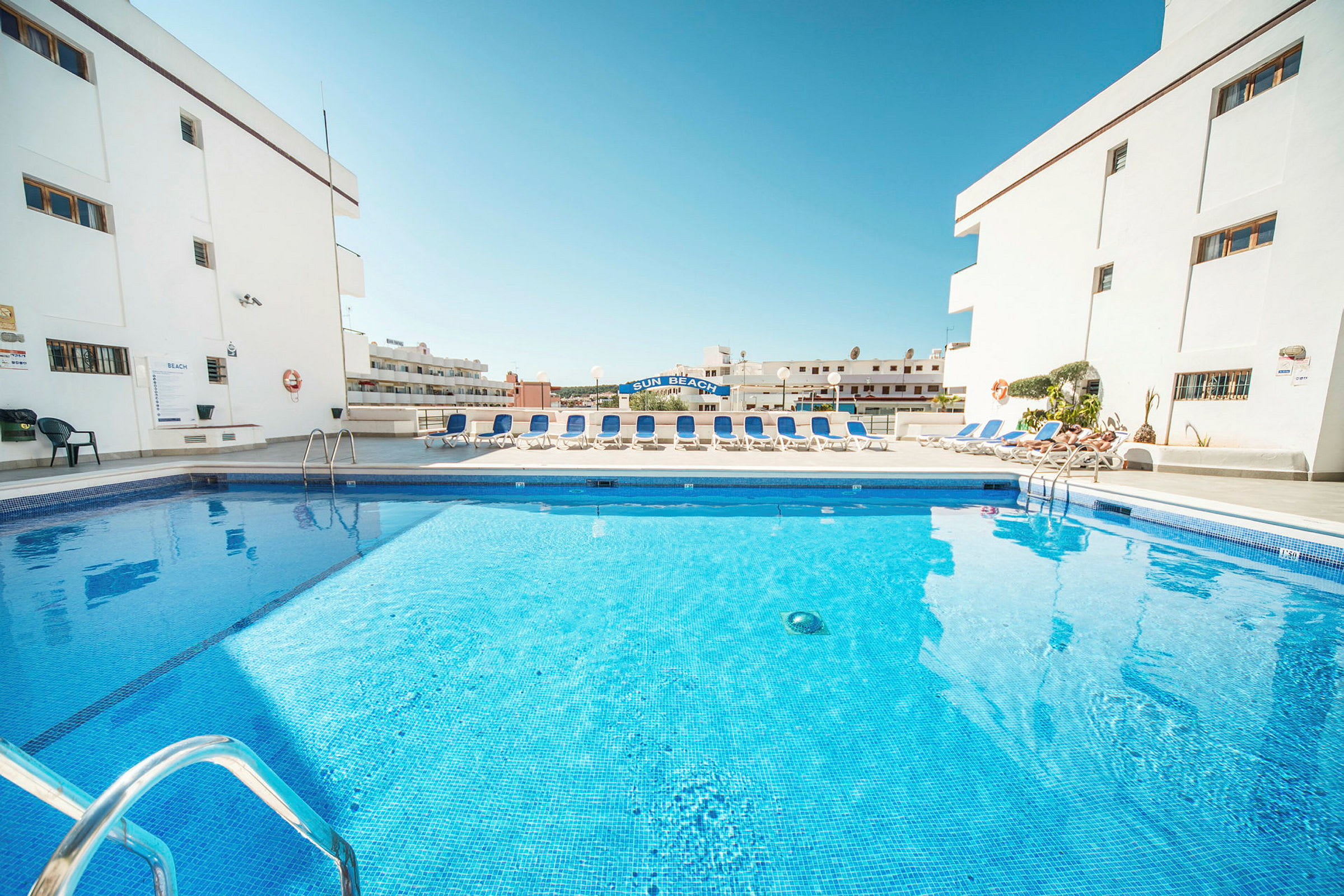 Bild av en swimmingpool omgiven av solstolar och vita byggnader. (AI-genererad text)