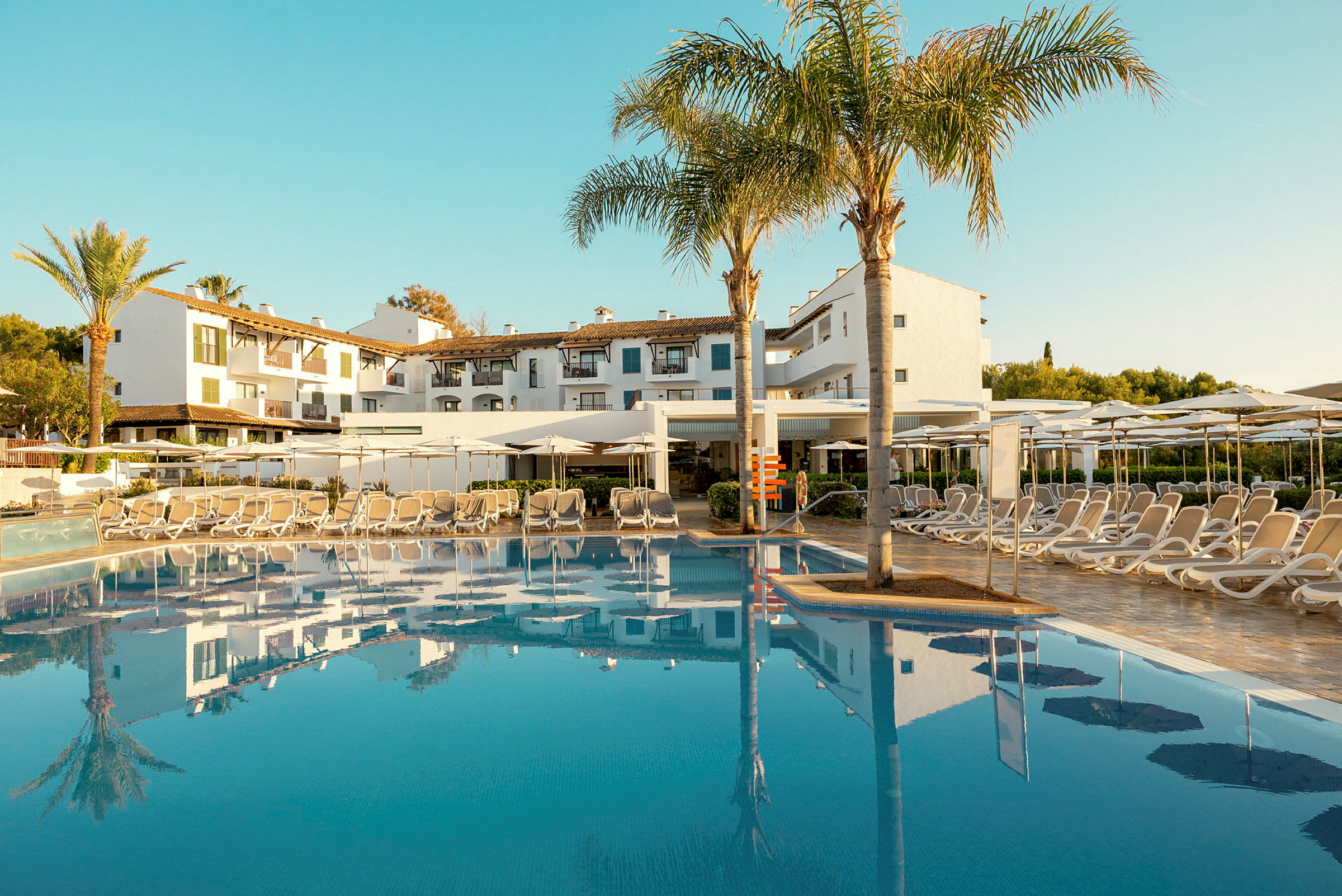 Hotellbyggnad bakom en pool med solstolar och parasoller. (AI-genererad text)