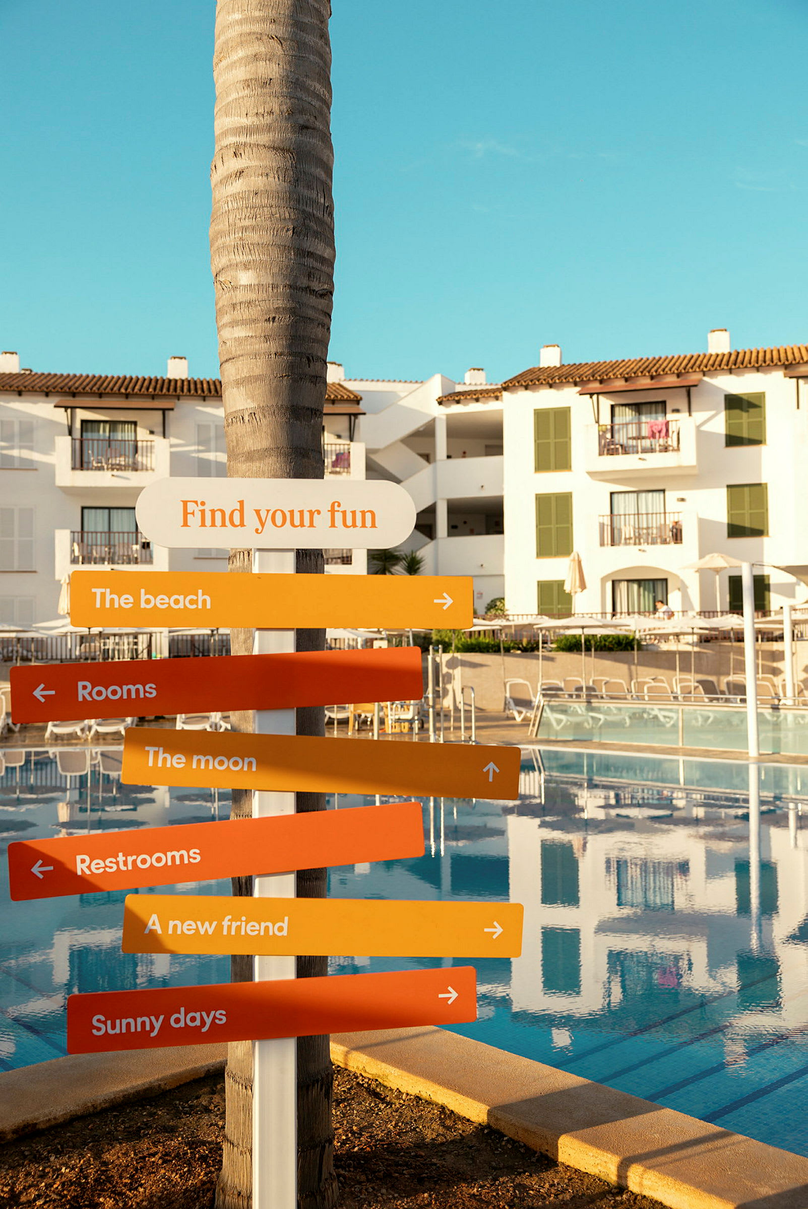 En skylt med texten 'Find your fun' och flera färgade pilar på en resort med pool och vita byggnader. (AI-genererad text)