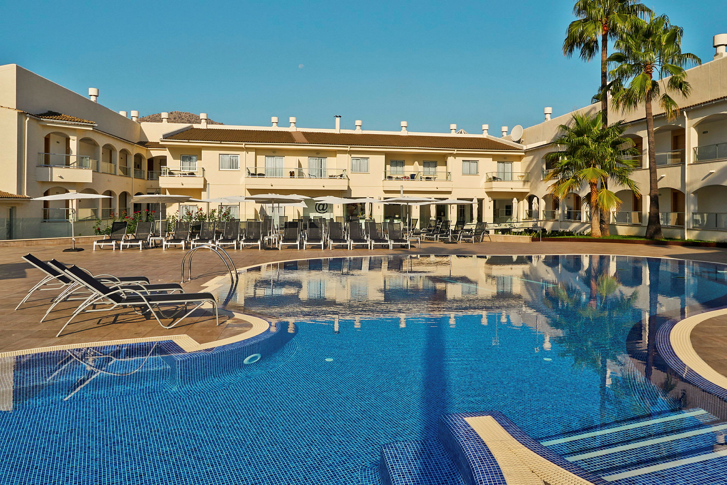 Hotellbyggnad med pool och solstolar i förgrunden. (AI-genererad text)