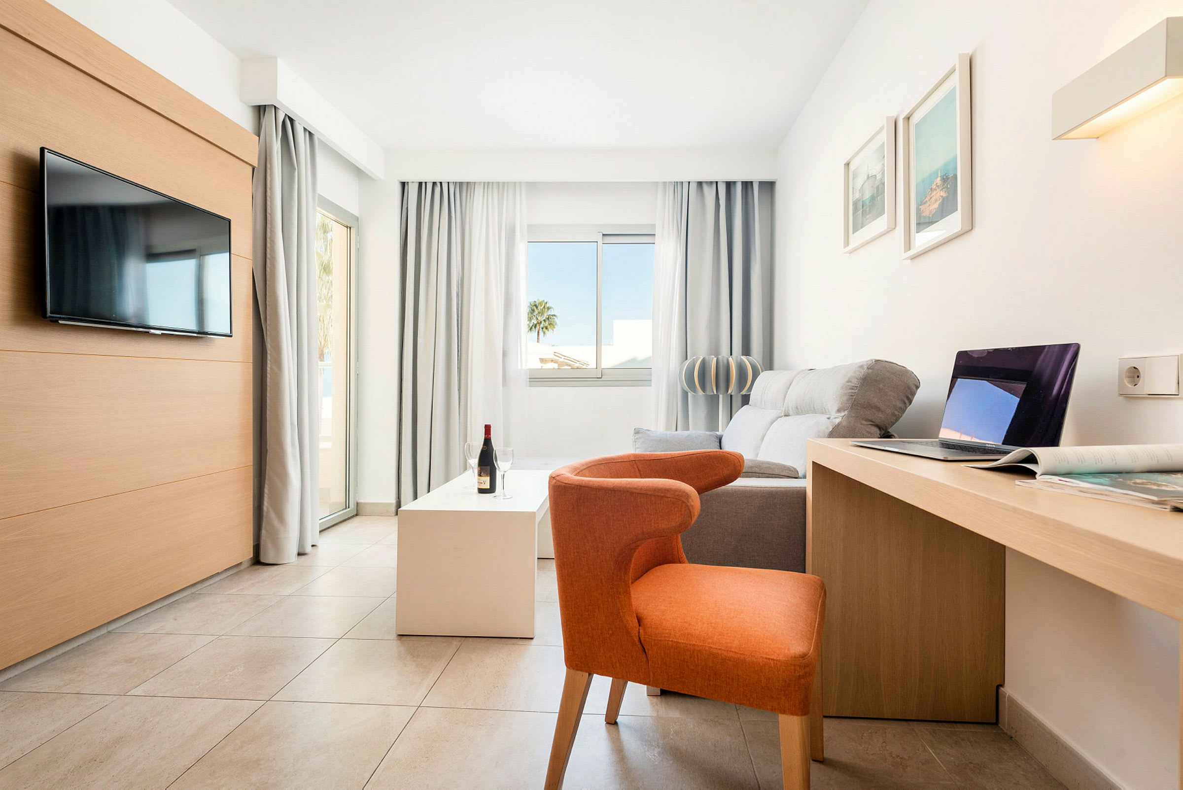 Ljust, modernt hotellrum med soffa, stol, TV, skrivbord och stora fönster. (AI-genererad text)