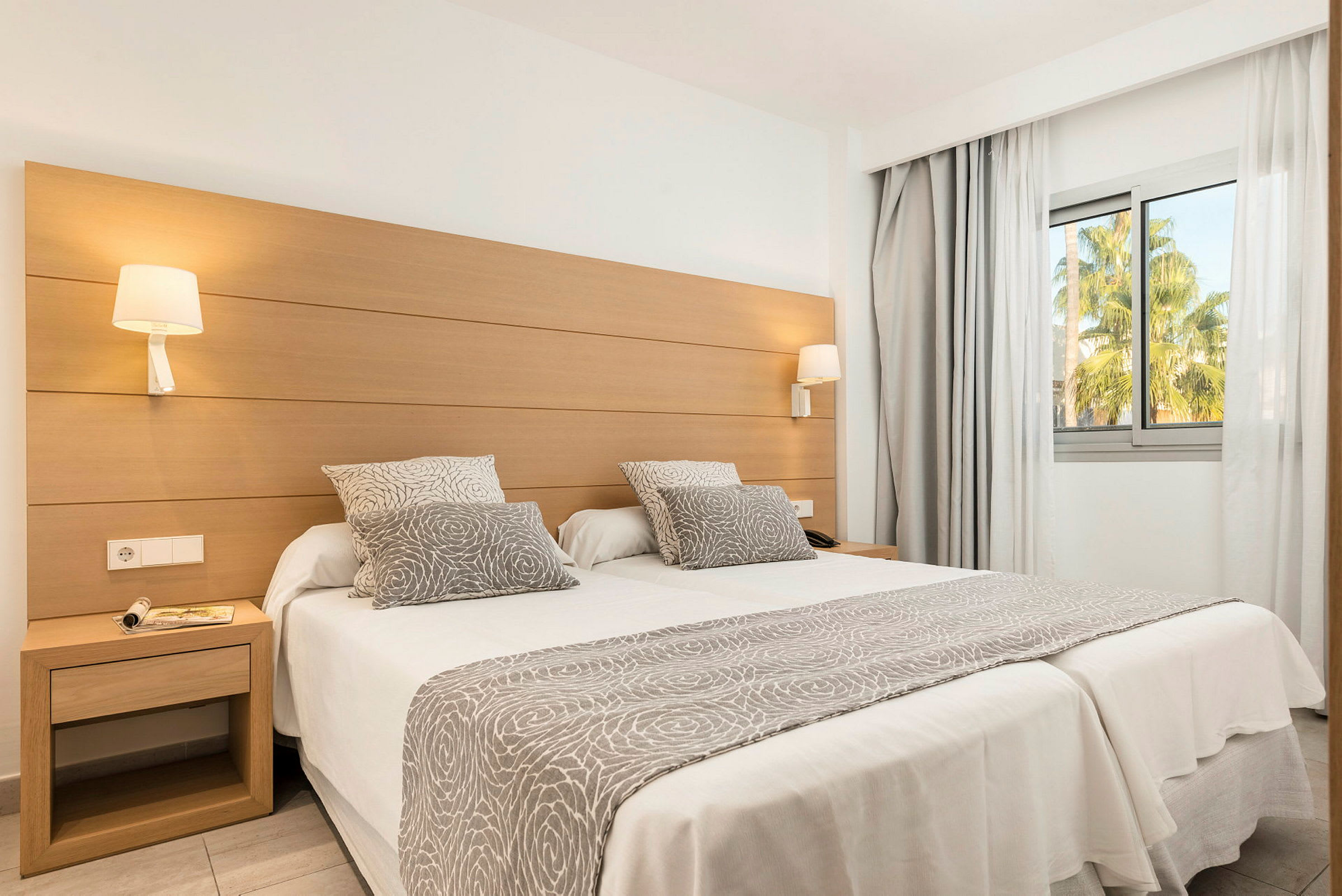 Ett hotellrum med två enkelsängar sammansatta, dekorerat med gråvita textilier med mönster, och ett stort fönster med utsikt mot grönska. (AI-genererad text)