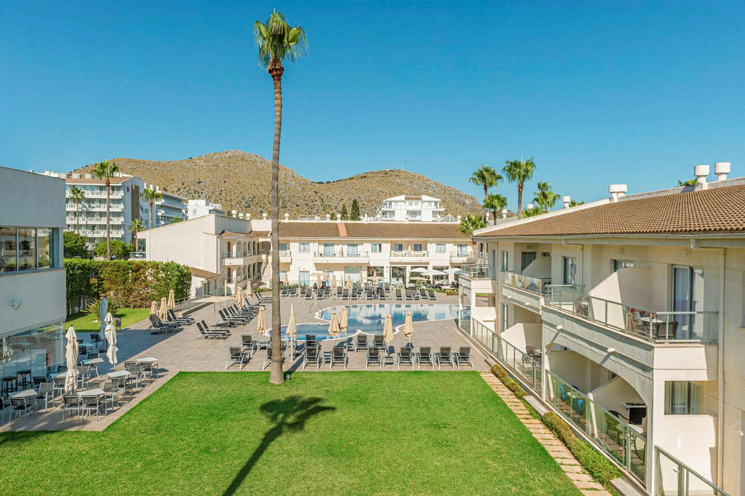 Vy över hotellområde med byggnader, pool och gräsmatta, omgiven av palmer och berg i bakgrunden. (AI-genererad text)