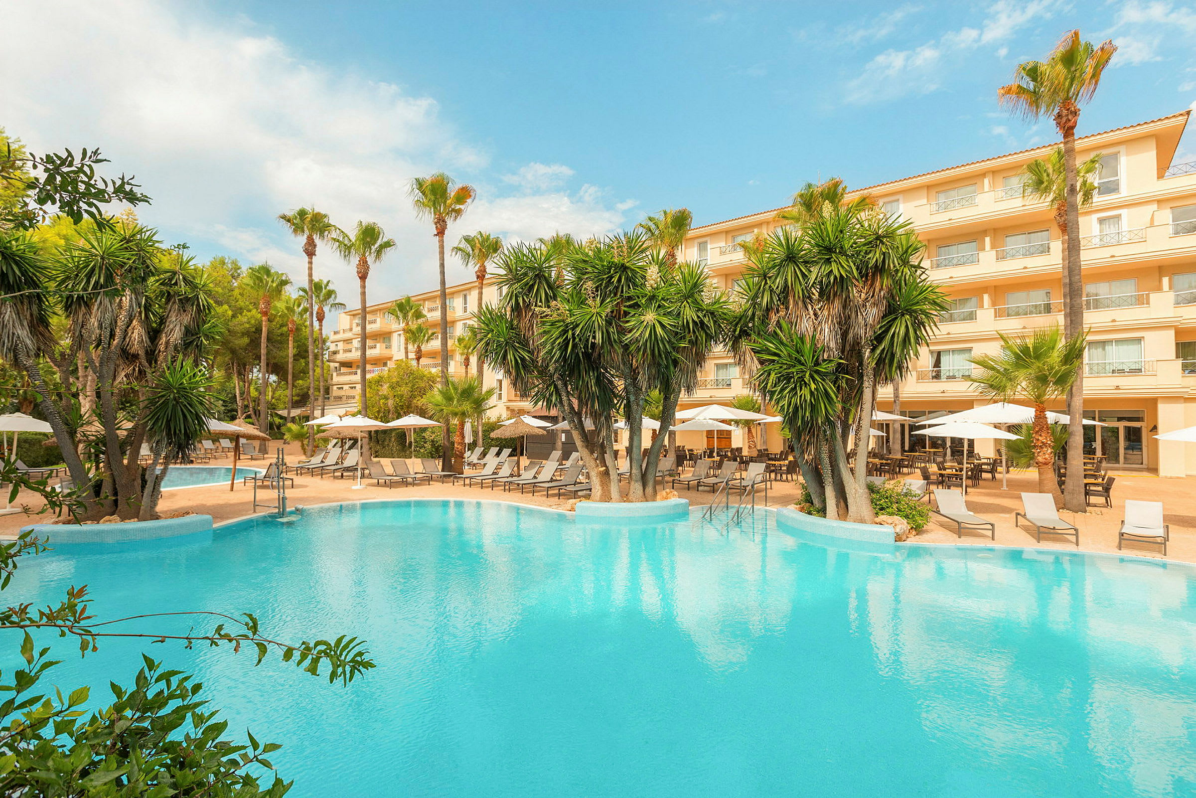 Hotellbyggnad med balkonger och möblerade uteplatser vid en pool omgiven av palmer och solstolar. (AI-genererad text)