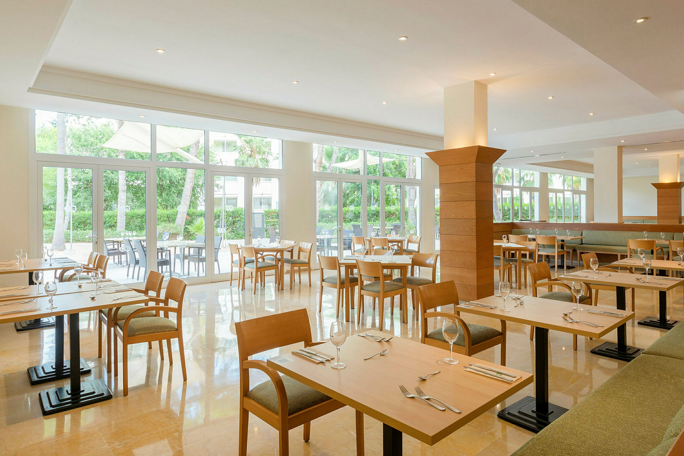 Ljus och rymlig restaurang med träbord och stolar, omgiven av stora fönster med utsikt över grönska. (AI-genererad text)