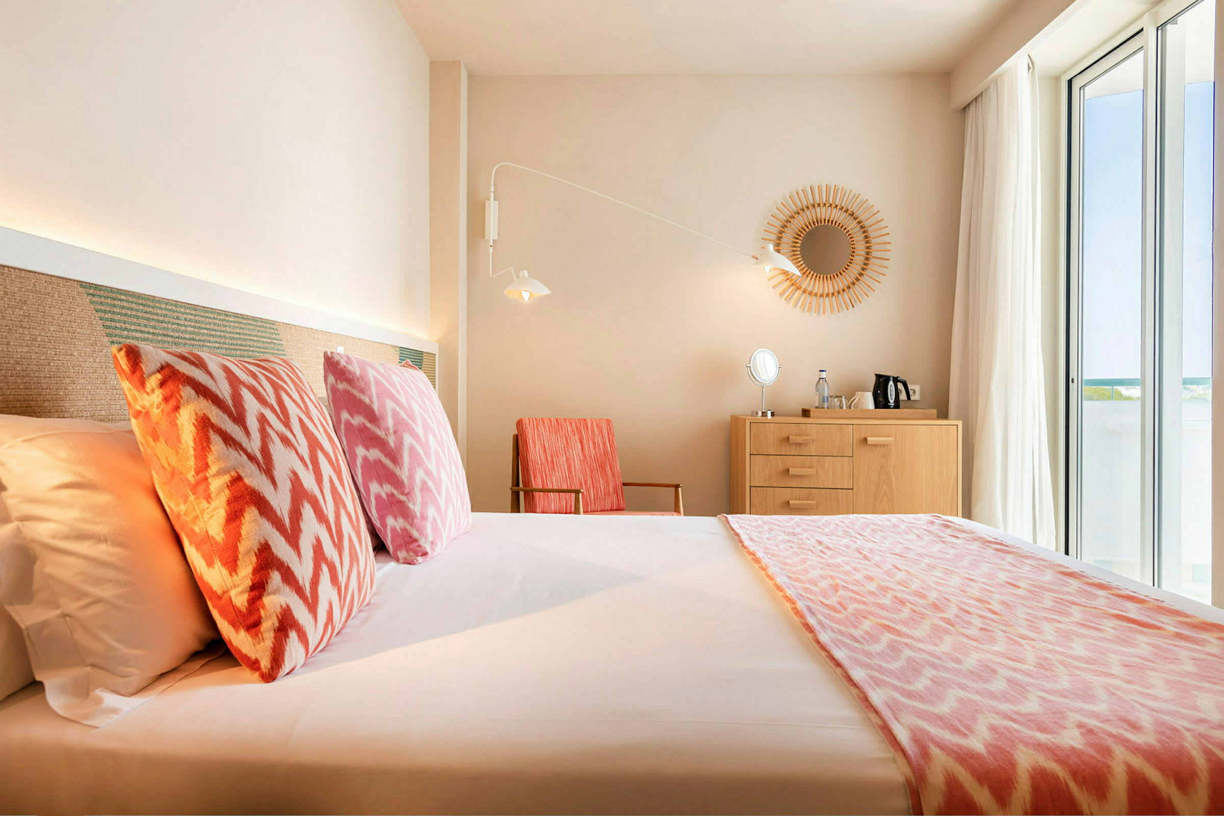 Ett hotellrum med en säng, färgglada kuddar, träkommod och stora fönster. (AI-genererad text)