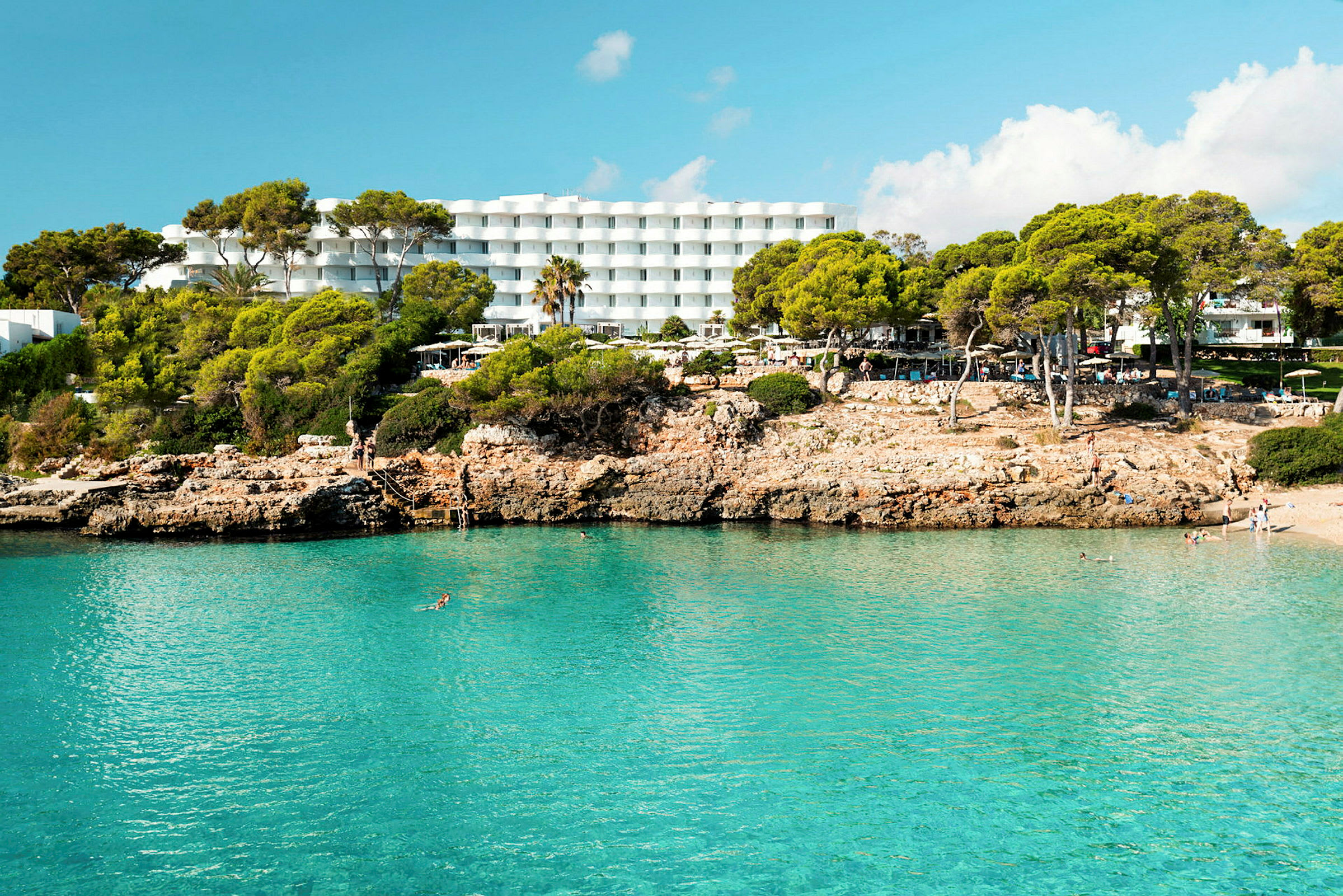 Hotell med vita byggnader bakom en klippig strand med klart blått vatten. (AI-genererad text)