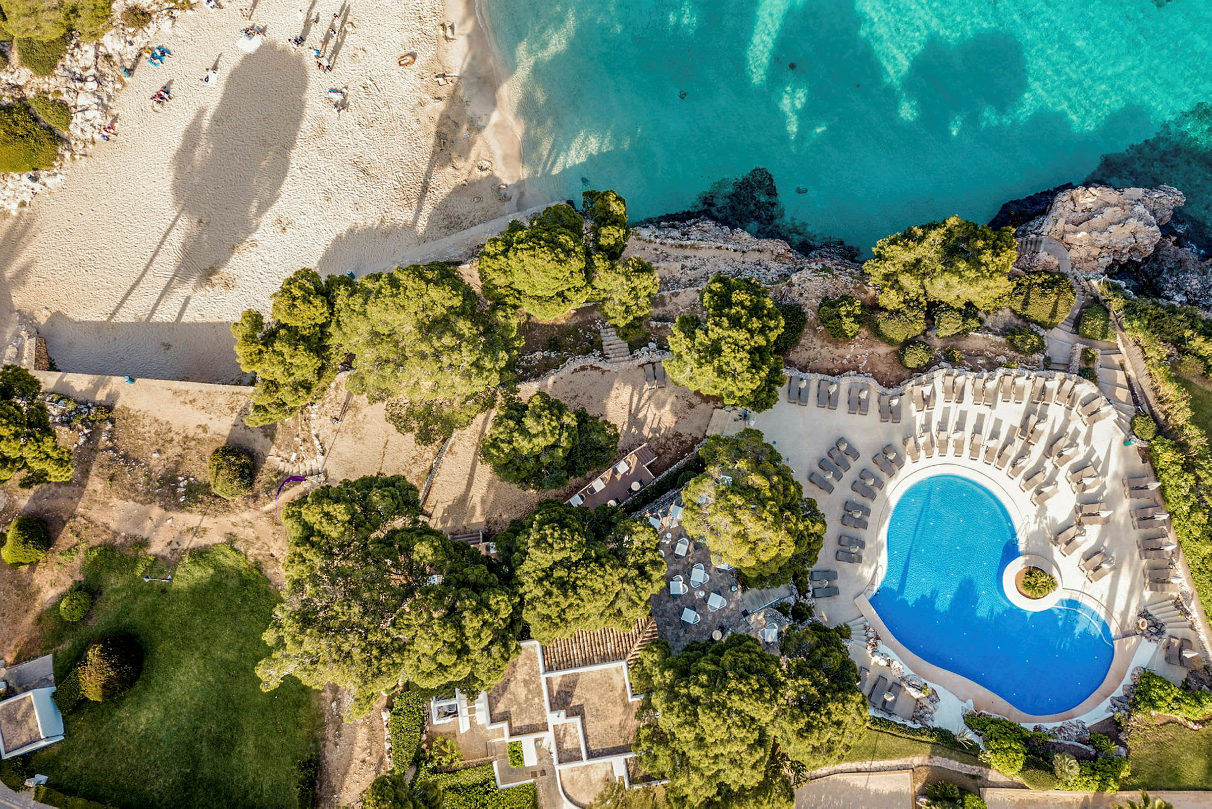 Flygfoto över en pool, omgiven av solstolar, med strand och turkost vatten i bakgrunden. (AI-genererad text)