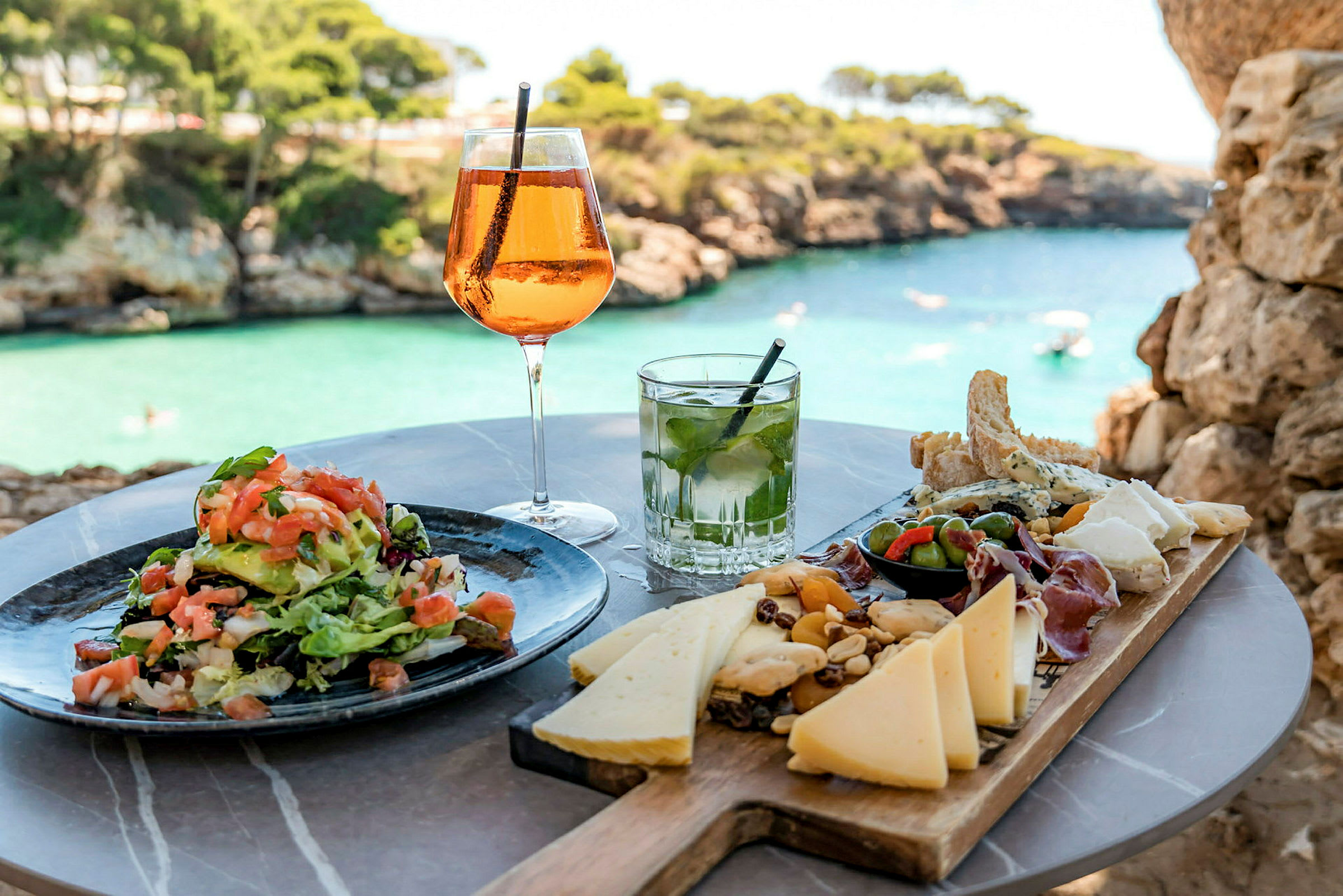 Ett bord med en sallad, ostar och drinkar med utsikt över vatten och klippor. (AI-genererad text)