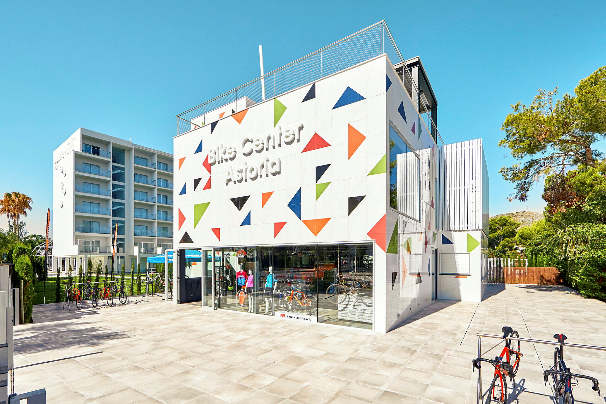 En modern byggnad dekorerad med färgglada triangulära mönster, märkta med texten "Bike Center Astoria". Framför byggnaden står cyklar och det är en klar blå himmel. (AI-genererad text)