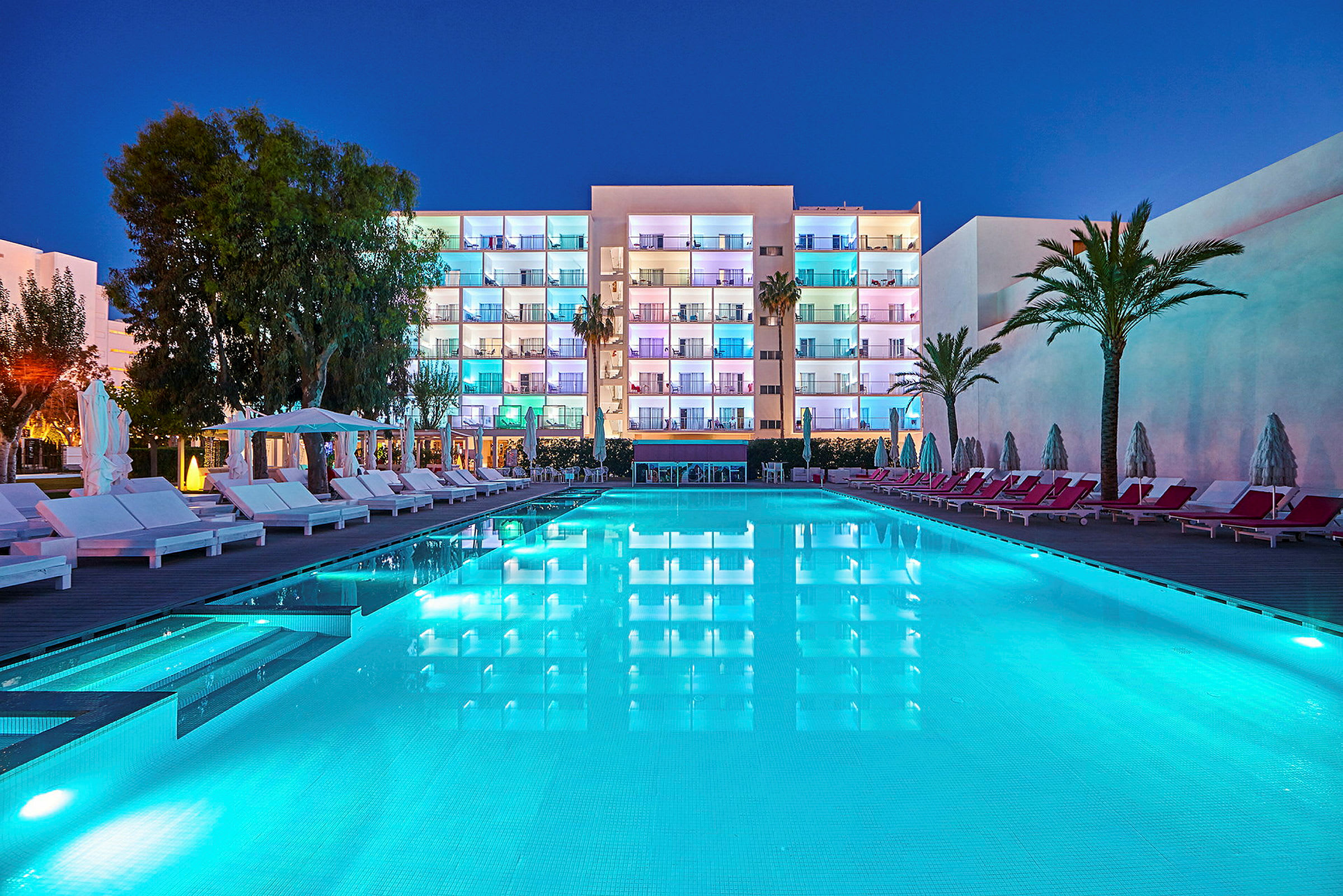 Bilden visar en belyst hotellbyggnad på kvällen med en pool i förgrunden omgiven av solstolar och parasoller. (AI-genererad text)