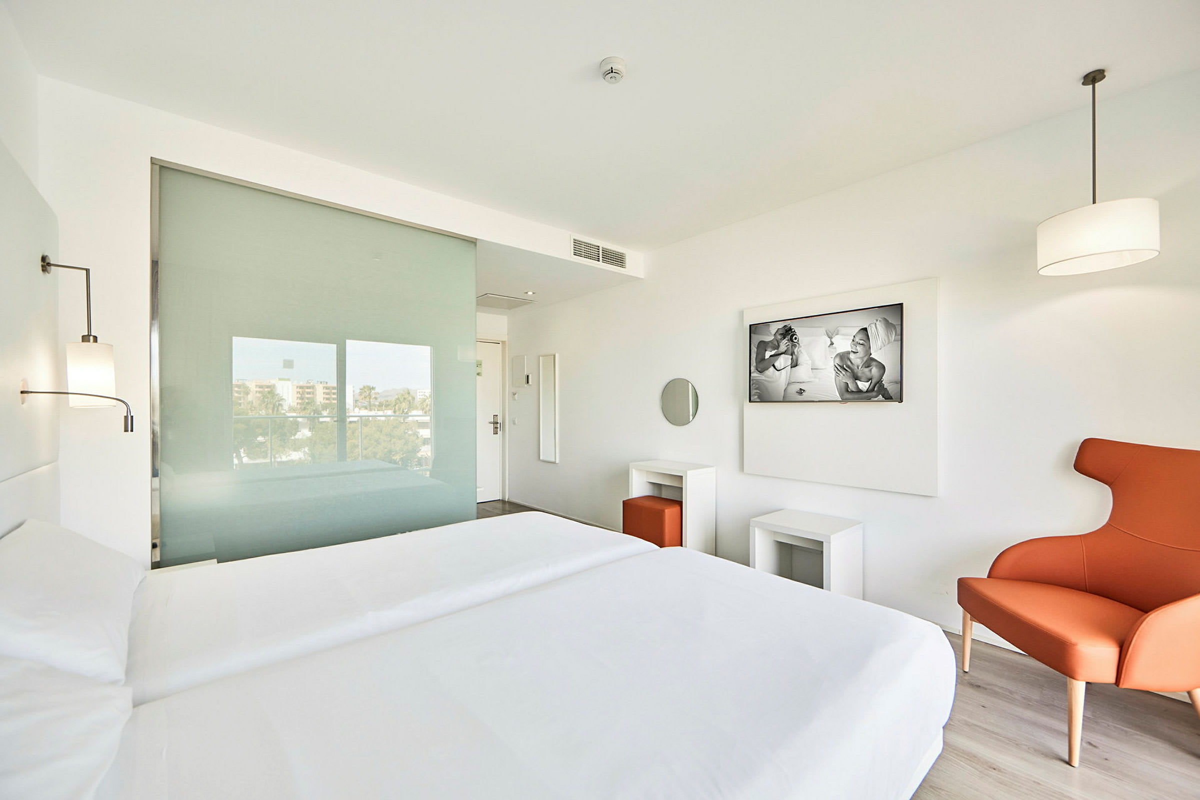 Bild av ett modernt inrett hotellrum med vita väggar, en dubbelbädd, en orange fåtölj och stora fönster. (AI-genererad text)