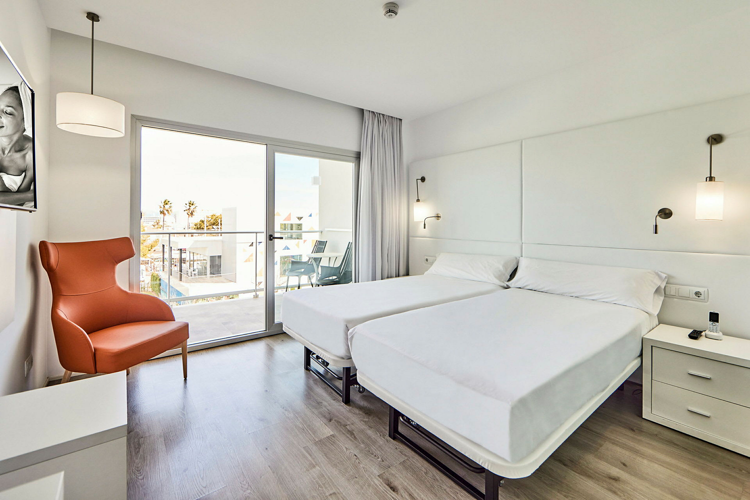 Ett ljust och modernt hotellrum med två enkelsängar, en orange fåtölj och ett glasfönster som leder till en balkong. (AI-genererad text)