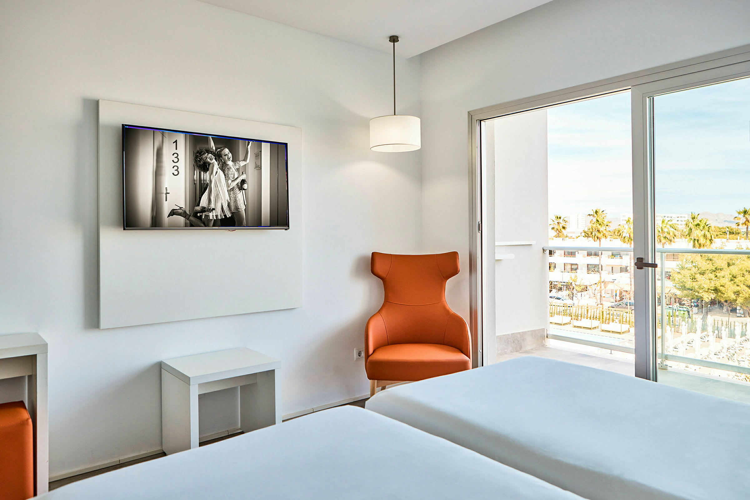 Ett hotellrum med två enkelsängar, en orange fåtölj och en TV på väggen. Genom ett fönster syns en balkong och utomhusmiljö. (AI-genererad text)
