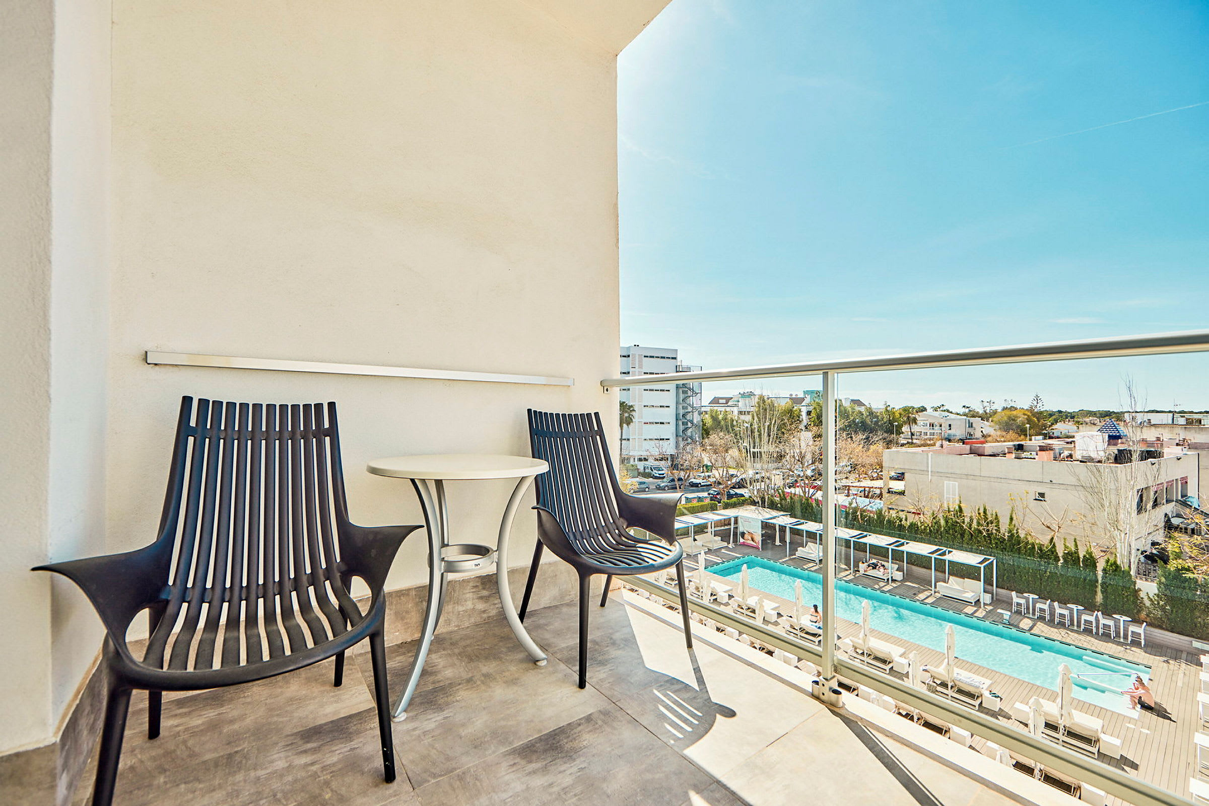 En balkong med två stolar och ett bord, med utsikt över en pool och omgivande byggnader. (AI-genererad text)