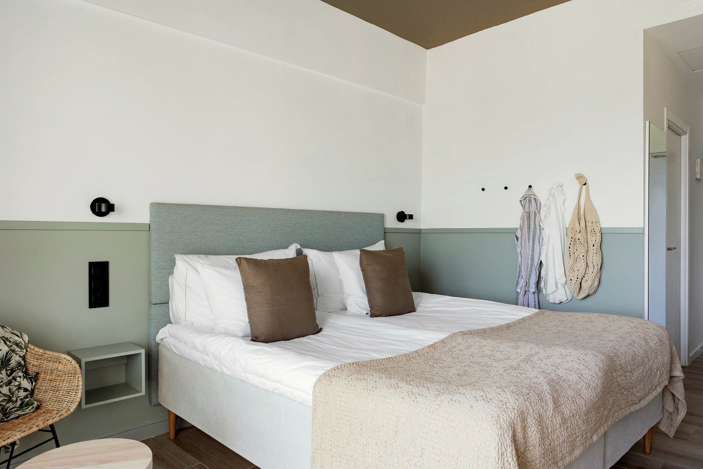 Ett hotellrum med en dubbelsäng, två kuddar och en beige överkast. Vägg med krokar och hängande klädesplagg. (AI-genererad text)