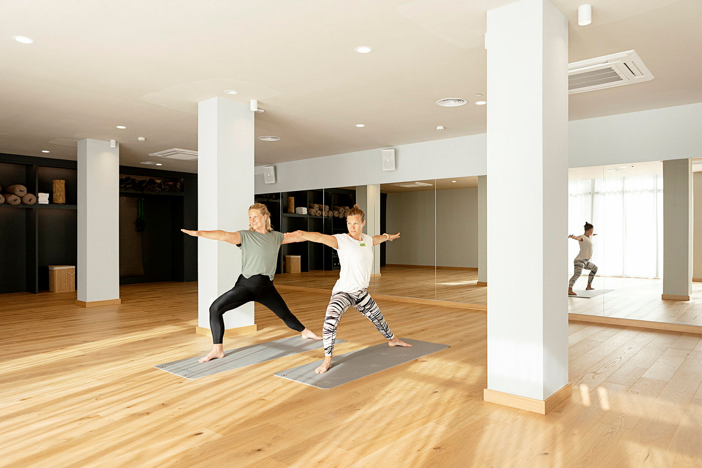 Två personer utför yoga i en ljus studio med trägolv och spegelvägg. (AI-genererad text)