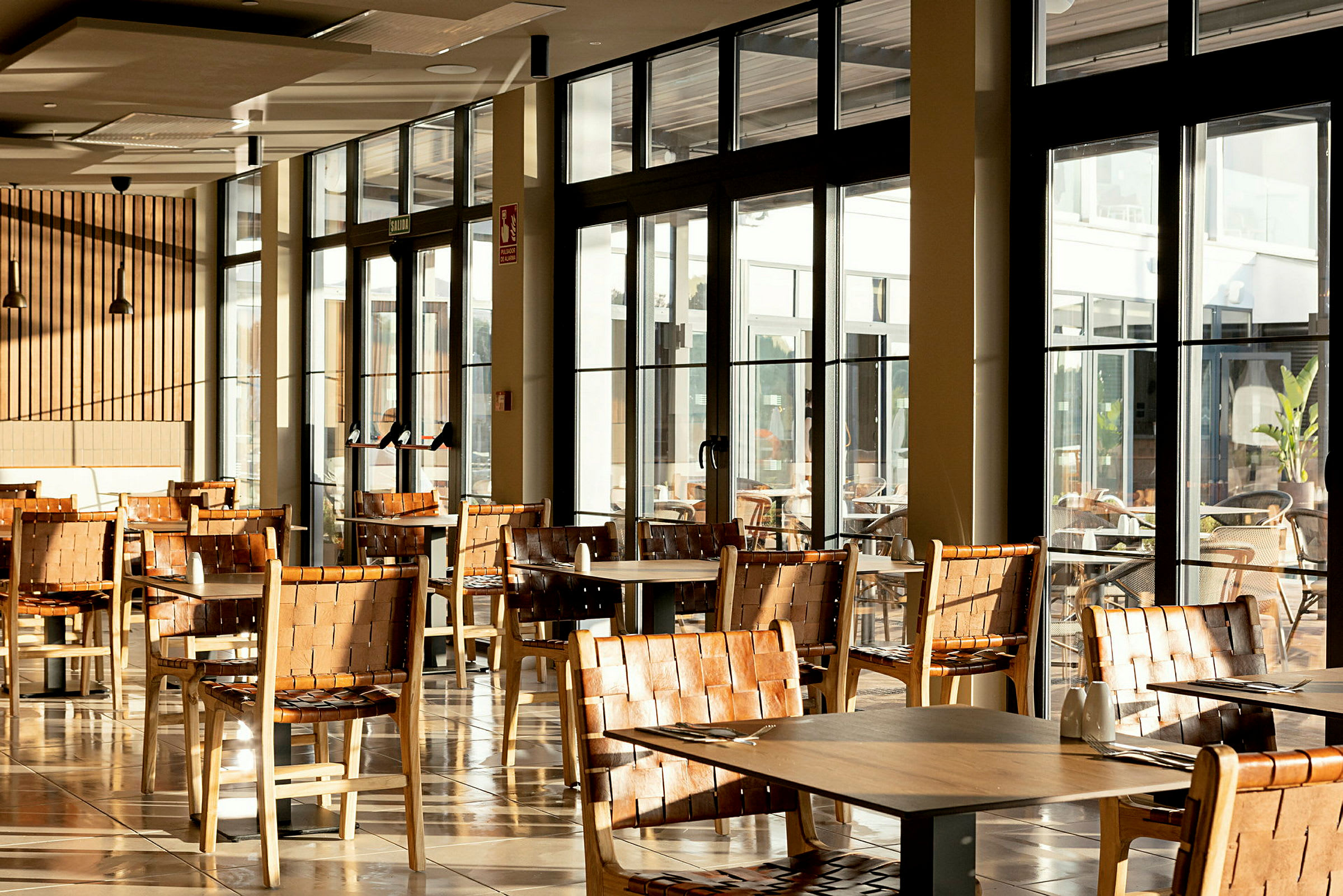 Restaurangområde med bord och stolar i trä, stora glasfönster med utsikt utåt. (AI-genererad text)