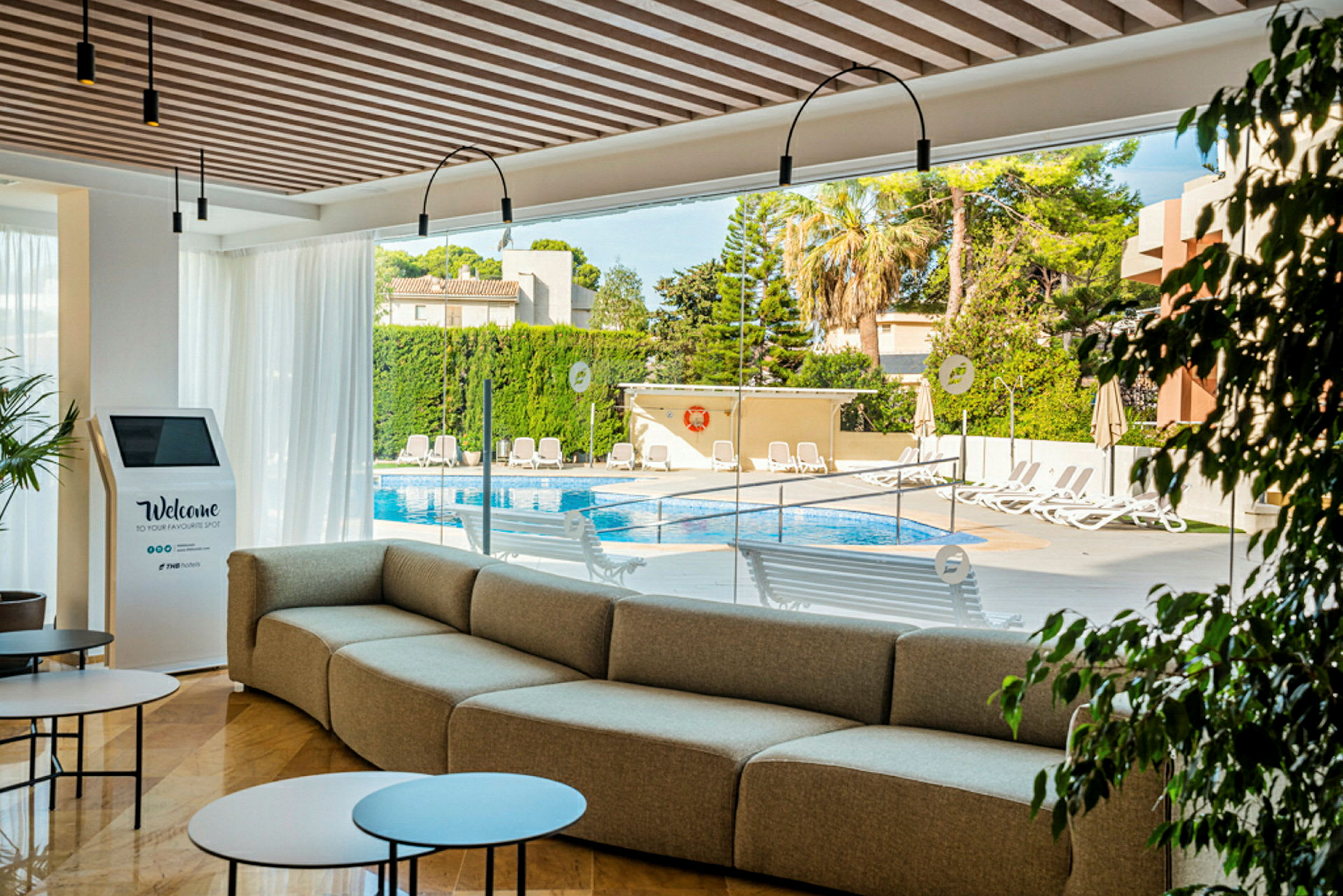 En modern lounge med soffa, bord och utsikt mot poolområdet utanför glasväggarna. (AI-genererad text)