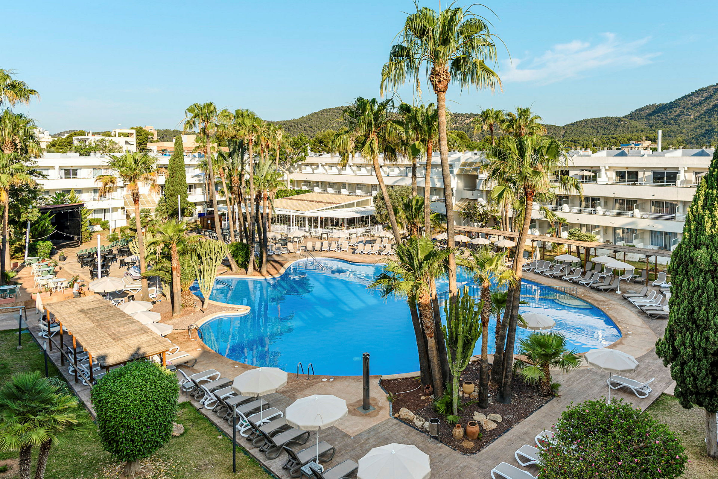 En hotellpool omgivet av solsängar och parasoller, med palmer och byggnader i bakgrunden. (AI-genererad text)