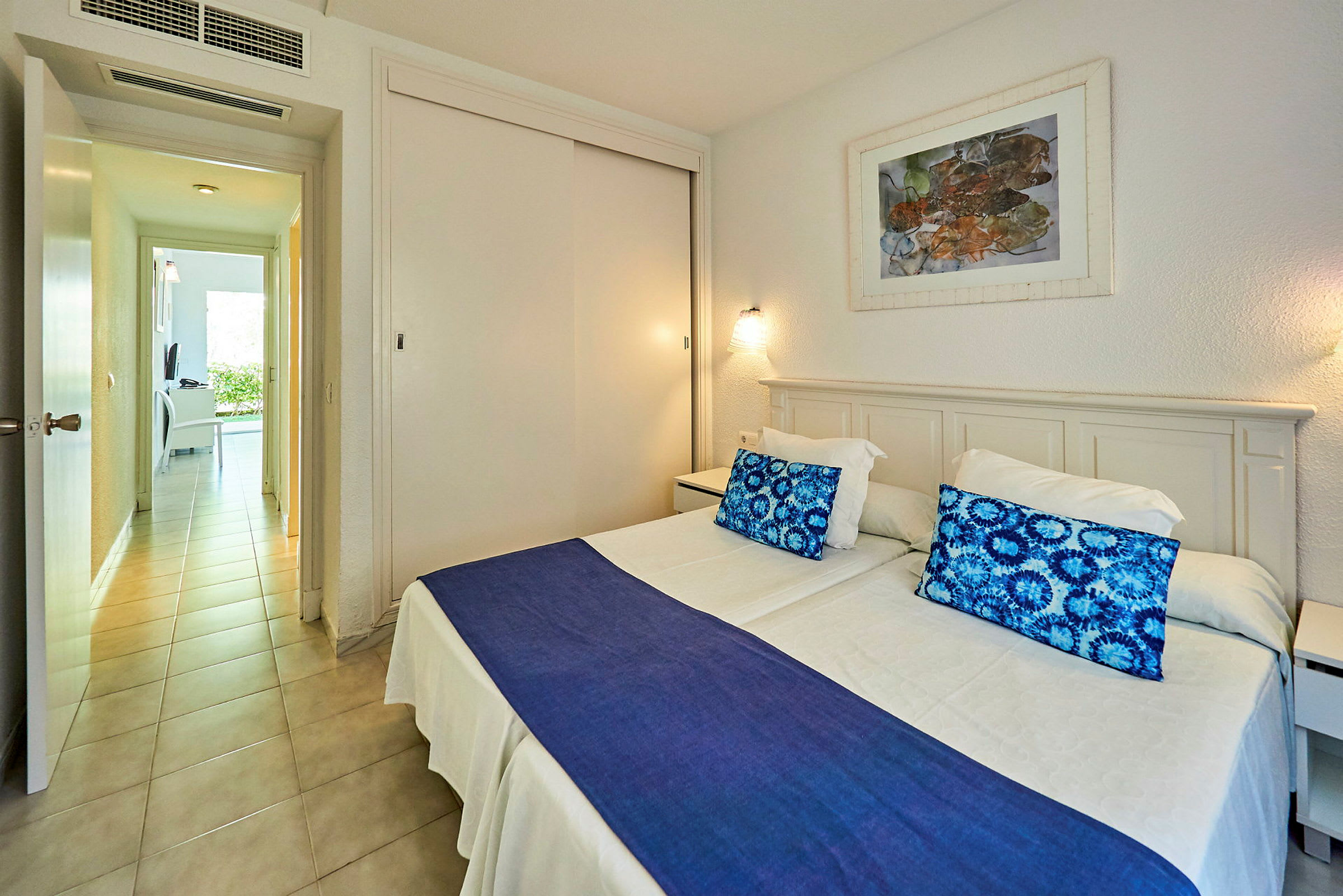 Ett hotellrum med två enkelsängar, färgglada kuddar och en blå överkast. (AI-genererad text)
