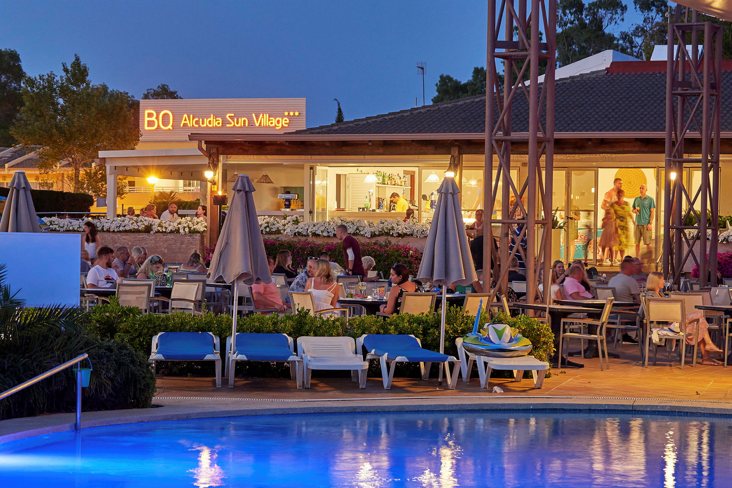 Uteservering vid ett hotell med gäster och en pool i förgrunden, kvällstid. (AI-genererad text)