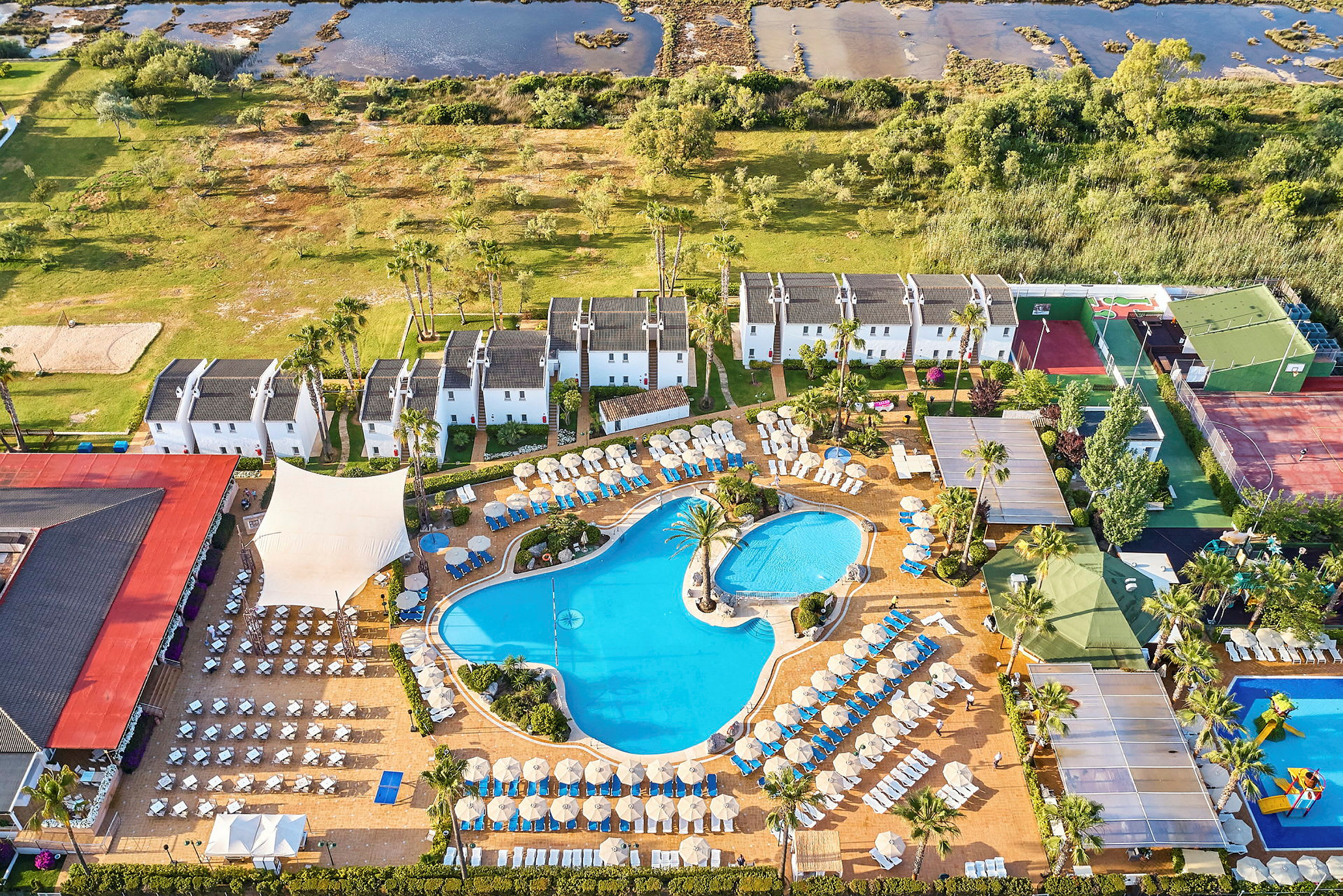 Flygvy över hotellområde med pooler, solstolar och vitmålade byggnader omgivet av grönska. (AI-genererad text)