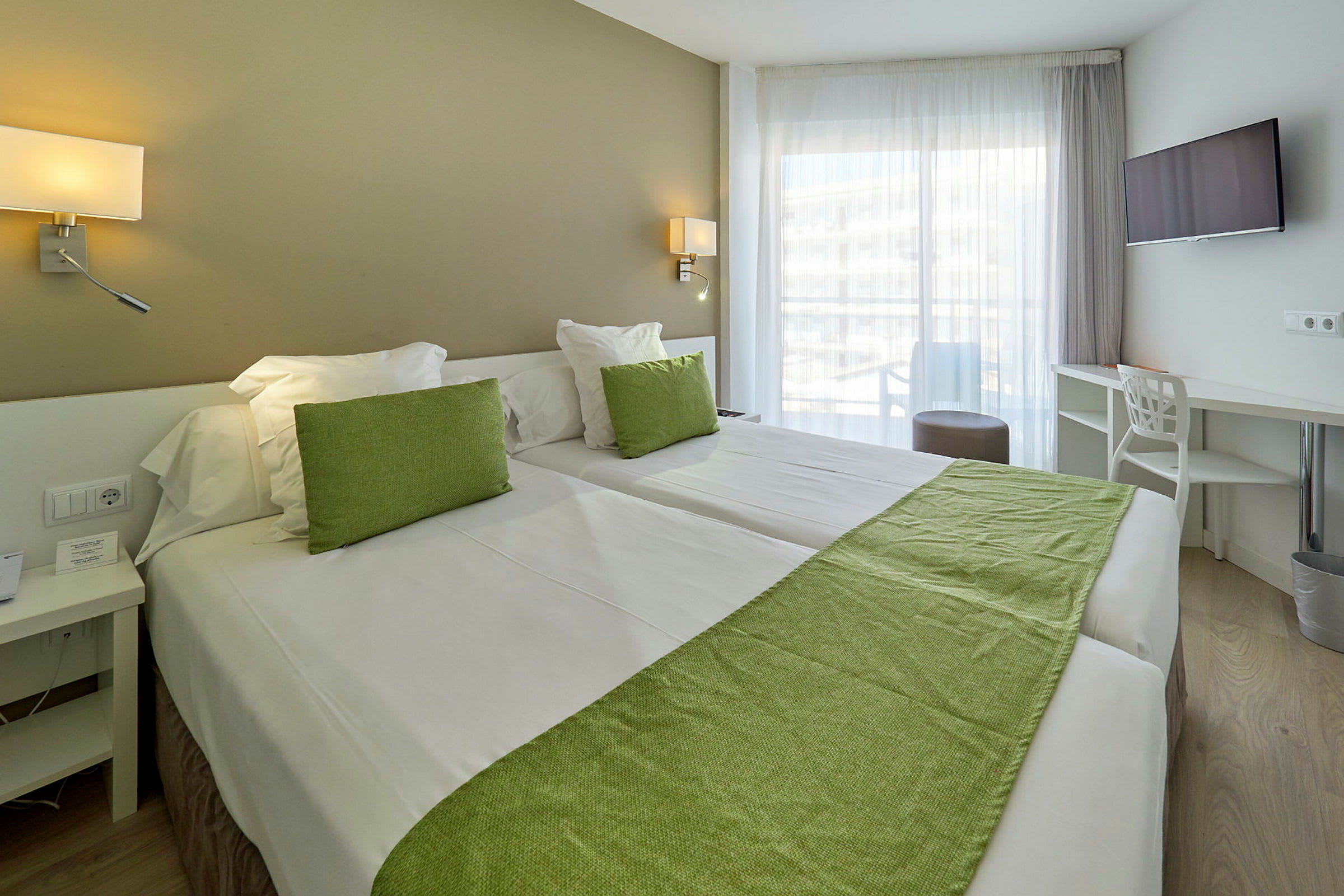 Hotellrum med två sängar, grön dekoration och stora fönster. (AI-genererad text)