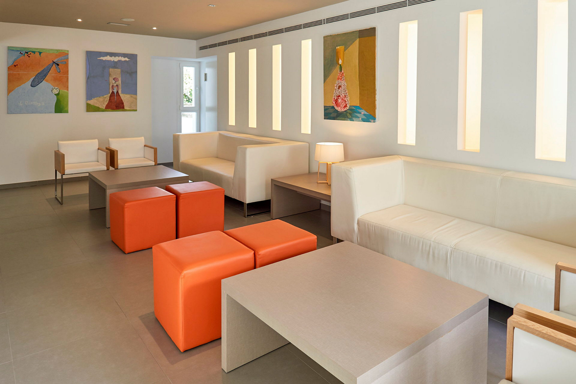 Ett modernt lobbyområde med vita soffor och orange sittpuffar, kompletterat med konstverk på väggarna. (AI-genererad text)