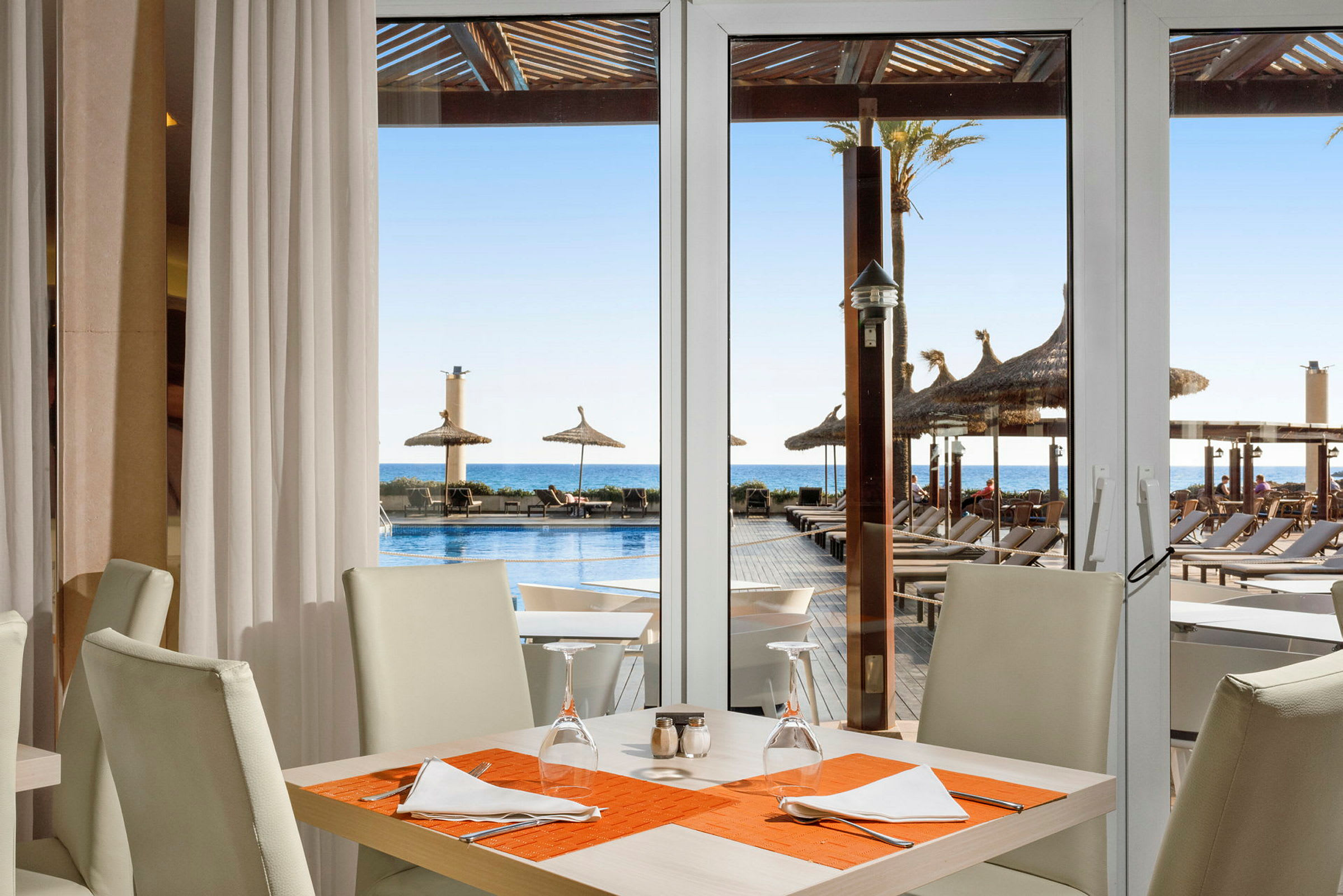 Ett uppdukat bord med vita stolar och orange bordsduk, med utsikt mot en pool och solstolar i bakgrunden. (AI-genererad text)