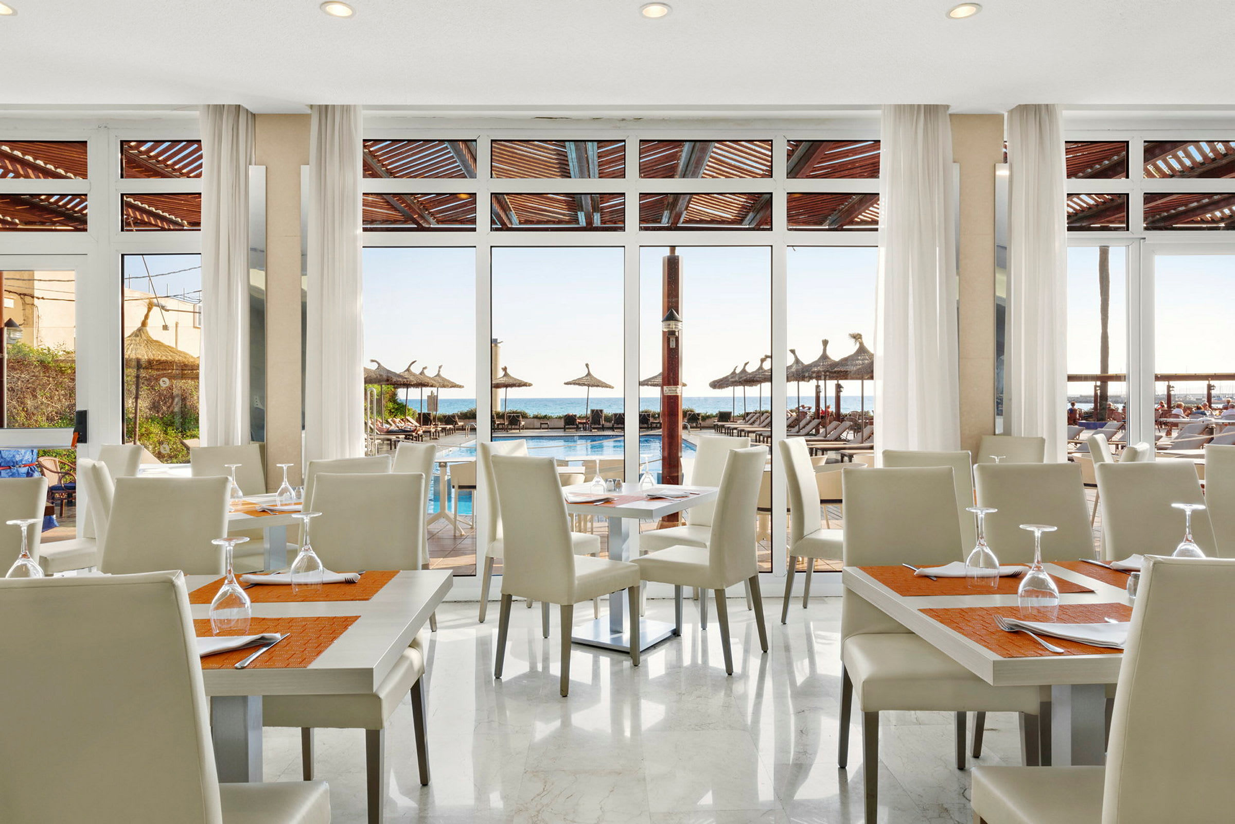 En restaurang med vita stolar och bord, med utsikt mot en pool och havet genom stora glasfönster. (AI-genererad text)
