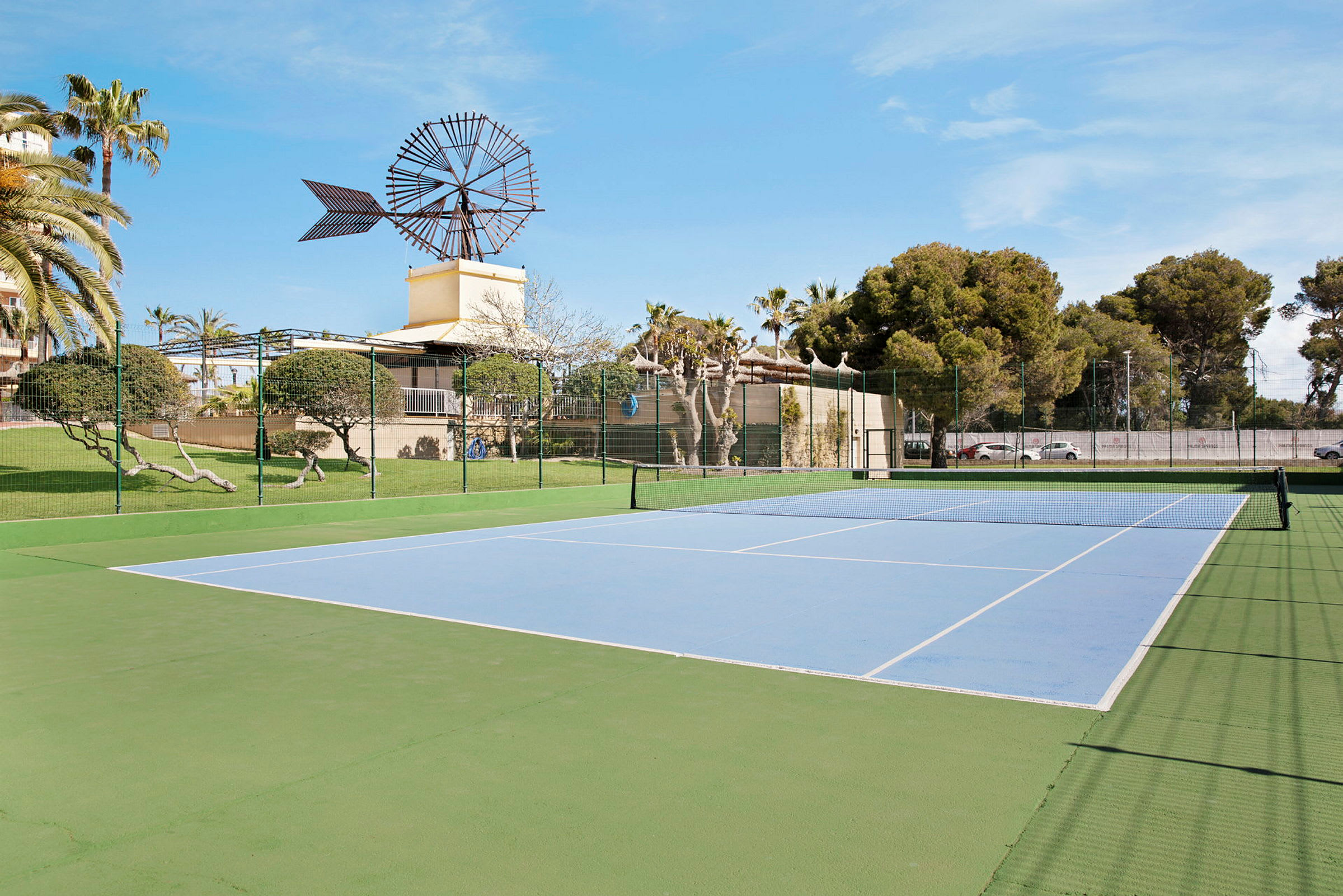 En utomhus tennisbana med en väderkvarn i bakgrunden och omgivande grönska. (AI-genererad text)
