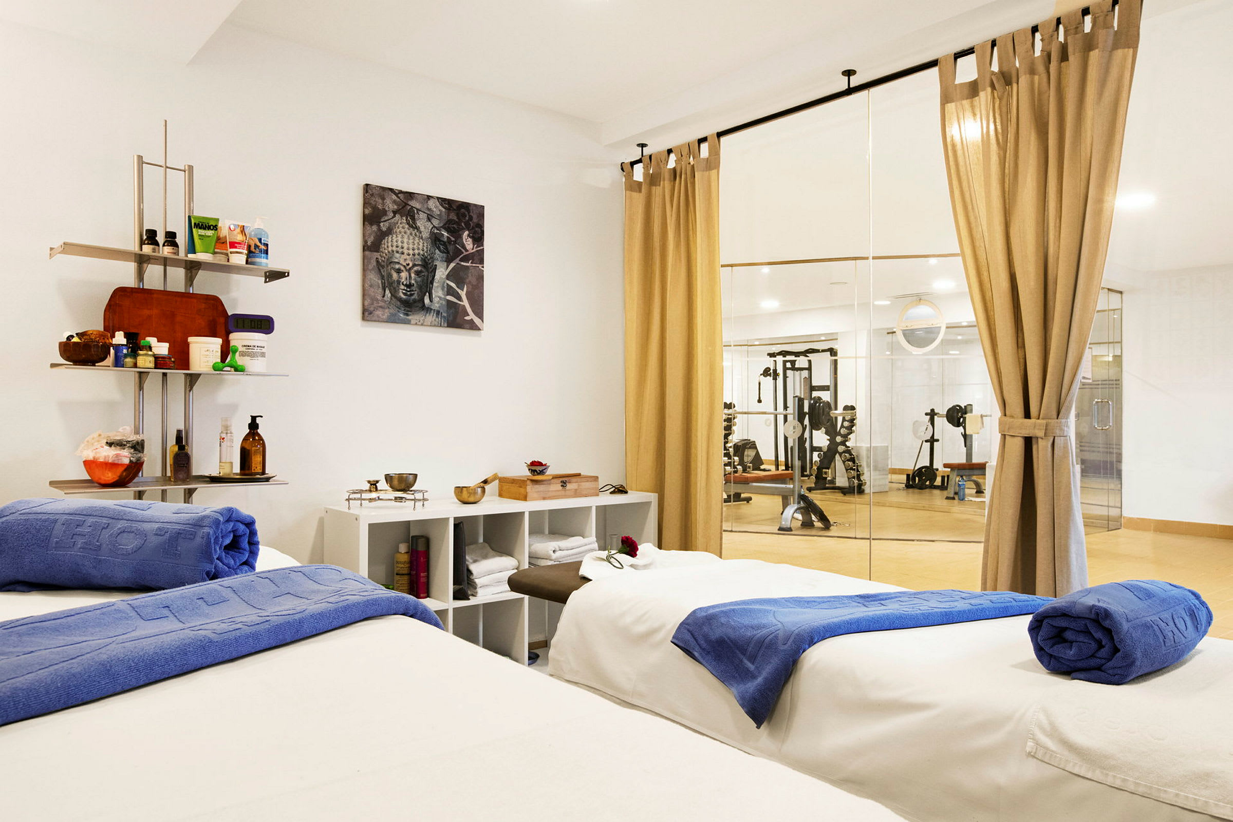 Interiör av spa-rum med två massagebänkar och träningsutrustning synlig genom glasvägg. (AI-genererad text)