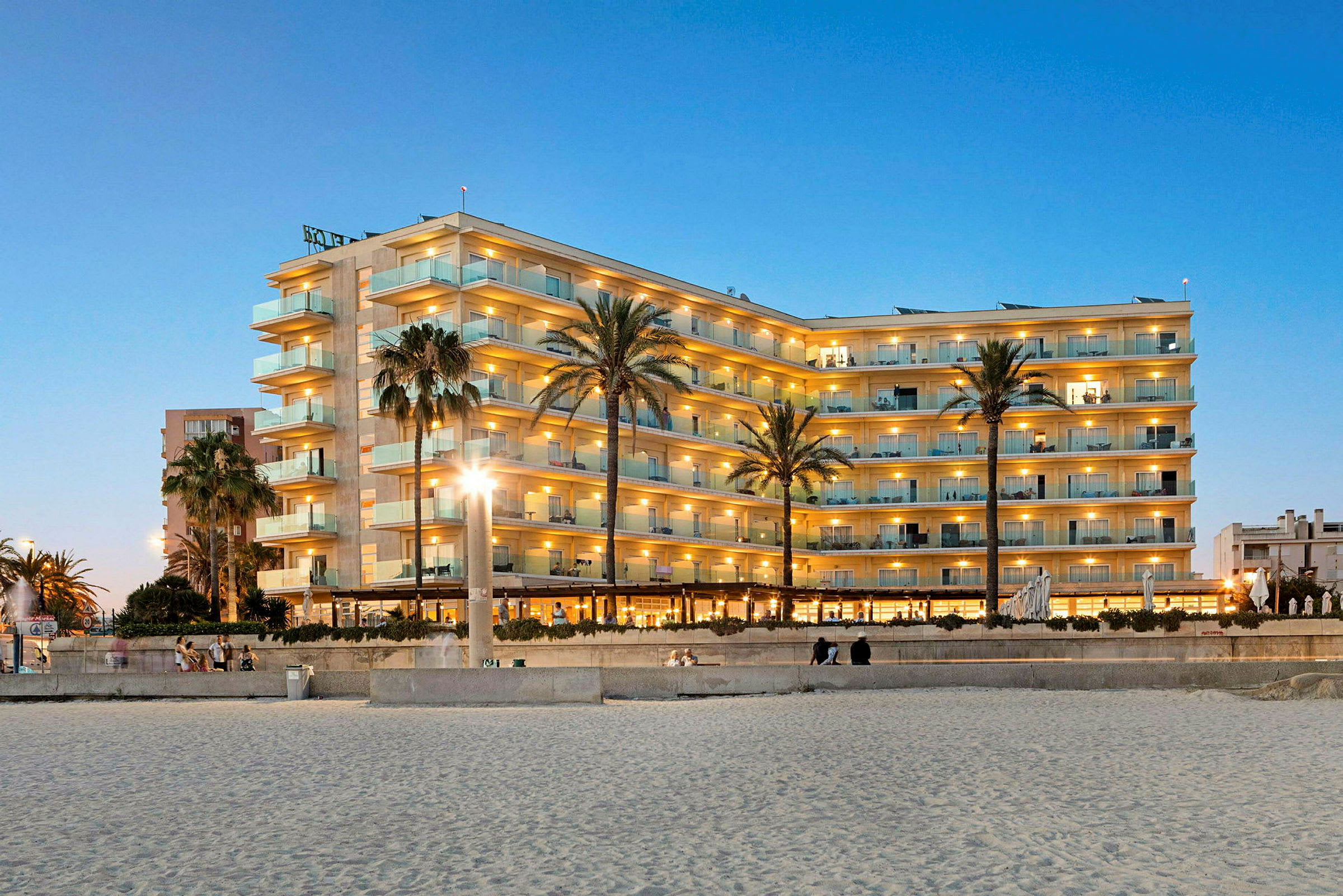 Hotellbyggnad med flera våningar, belägen nära stranden, upplyst på kvällstid med palmer i förgrunden. (AI-genererad text)