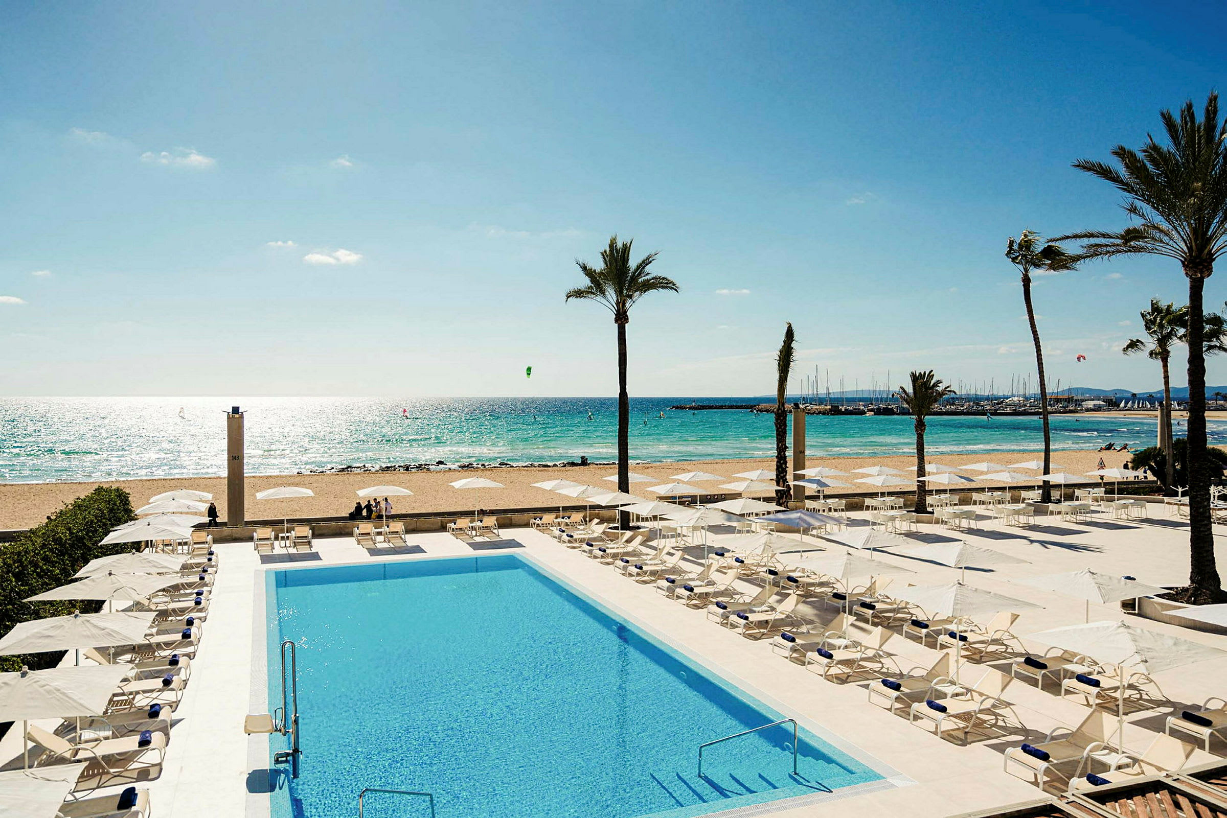 Pool med solstolar, palmer och strand med hav i bakgrunden. (AI-genererad text)
